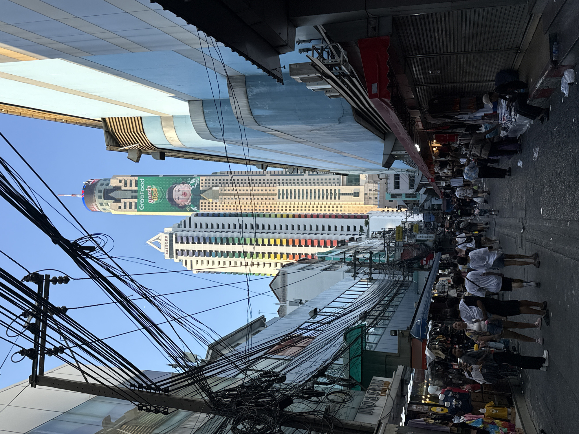 The image depicts a street scene in Bangkok, Thailand. Overhead, a dense network of tangled electrical wires dominates the view. On the right side of the frame, a crowded sidewalk lined with vendors selling goods is visible. Pedestrians, mostly dressed in casual attire, walk along the street. In the distance, tall buildings rise, including one adorned with a large "GrabFood" advertisement, indicating the presence of the Baiyoke Tower II. The sky is clear and blue. The environment is urban and bustling.