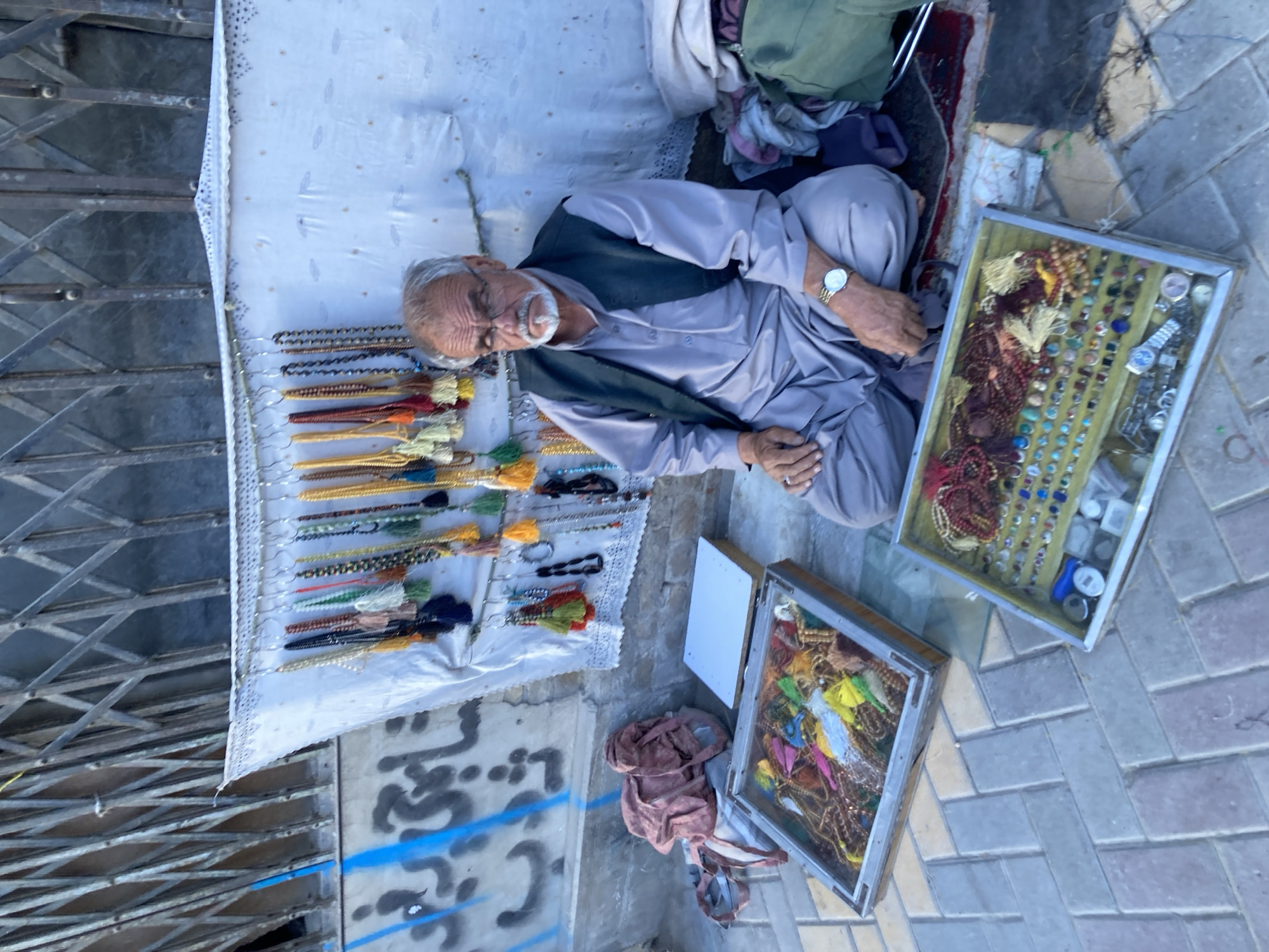 An elderly man with a white beard and glasses sits on a sidewalk in Quetta City Tehsil, Pakistan, next to his wares. He wears a light gray shalwar kameez and a dark vest. A white cloth spread out on the ground displays rows of prayer beads and jewelry. Two framed trays on the ground contain more beads, stones, and watches. The man sits cross-legged, his hands resting on his lap. A metal gate stands to his left, and graffiti marks the wall behind him. The sidewalk consists of light-colored bricks.