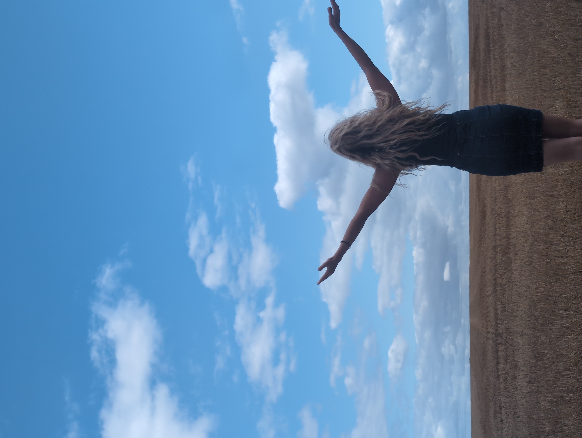 A person with long, blonde, wavy hair is seen from behind, standing in a field of dry, golden vegetation. The person is wearing a dark-colored dress or skirt and has their arms outstretched upwards and outwards. The background consists of a bright blue sky with scattered white clouds. The horizon line where the field meets the sky is visible. The image is taken outdoors, likely in a rural or agricultural setting, and the lighting suggests it is daytime. The specific location in Cavron-Saint-Martin, France is not discernible from the visual information provided in the image.
