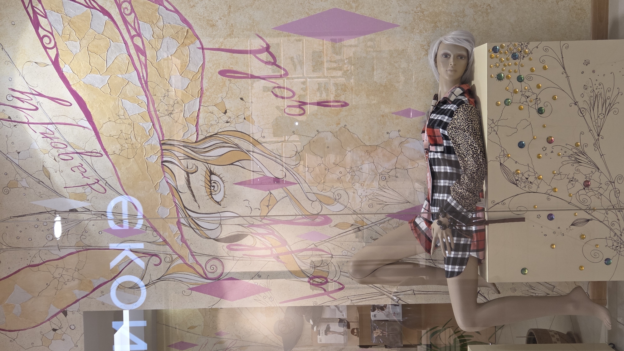 The image shows a mannequin lying on top of a decorated cabinet in what appears to be a shop window display. The mannequin has light-colored hair and is wearing a black and white plaid dress with leopard print accents. The cabinet is light brown and adorned with swirling line art and colorful gem-like embellishments.
Behind the mannequin, a large illustration is visible on the glass. This illustration features a stylized drawing of a woman's face, with large eyes and flowing hair. Above the woman's head, large wings resembling those of a dragonfly are depicted, with the text "dragon fly" written in a script font. The word "gold" is also visible in a similar script, positioned to the right of the illustration.
On the left side of the glass, the reflective surface shows part of the interior of a store, including what appears to be signage with Cyrillic letters, possibly reading "ЕКО" or similar. The background of the illustration has a textured, marbled appearance in shades of beige and pink.
The overall scene suggests a fashion display with a fantasy or whimsical theme. The location is identified as Novotlitsy, Russia, implying this is a storefront in that city.