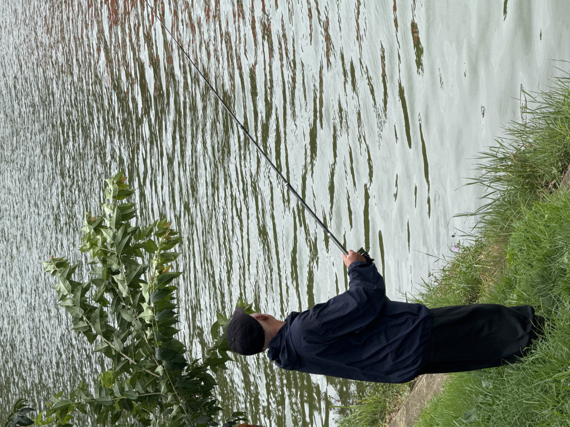 A man in a dark jacket and cap is fishing by the edge of a lake. He stands on a grassy bank, holding a fishing rod that extends into the rippling water. The lake is surrounded by lush green vegetation, with tree branches hanging over the water in the upper left corner. The scene appears to be a tranquil natural setting within the city of Da Lat, Vietnam.