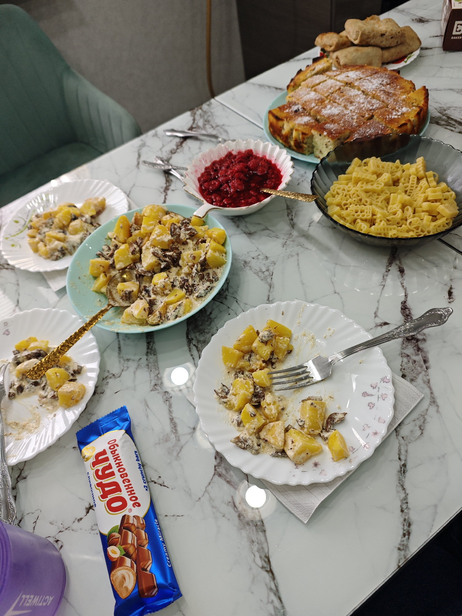 The image captures a casual meal set on a white marble-patterned table. In the foreground, several plates hold what appears to be a creamy potato and mushroom dish. A large light blue serving bowl in the center also contains this dish, with a golden serving spoon resting within. To the right, a plate with a silver fork has been partially eaten.
Further into the frame, a round cake or pie, dusted with powdered sugar and cut into slices, sits on a light blue plate. Behind it, a plate with several rolled pastries or blini is visible. Adjacent to the cake are two bowls: one filled with bright red berries, likely raspberries or cranberries, and another with alphabet-shaped pasta.
Other items on the table include a blue chocolate bar wrapper prominently featuring the brand "Обыкновенное Чудо" (Obyknovennoye Chudo, meaning "Ordinary Miracle"), along with text in English "x2 crunchy bars" and "Creamy Miracle". A purple cup, partially visible and inverted, displays the text "ACTIWELL". Part of a teal-colored upholstered chair is seen in the upper left background, and a brown box with "BAKER'S" partially visible in the upper right. The scene suggests a homemade meal or gathering, possibly in Фоки, Russia, given the brand names.