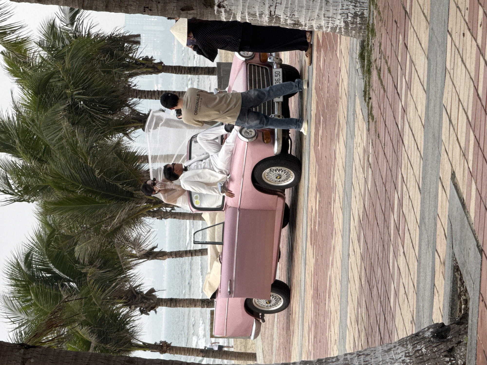A daytime outdoor scene unfolds on a paved promenade adjacent to a sandy beach in Đà Nẵng, Vietnam. The central focus is a photoshoot featuring a vintage pink two-door convertible car, equipped with spoked wheels and displaying the license plate "36.50."
A male subject, dressed in a white suit, black bow tie, and sunglasses, is seated on the car's hood, facing right. A female subject, wearing a white wedding gown, veil, sunglasses, and white gloves, is positioned within the car's open passenger doorway, her left arm resting on the vehicle's side.
Standing directly in front of the car, a photographer, wearing a beige t-shirt with "ORIGINALS VIETNAM" on the back, blue jeans, and a dark baseball cap, operates a camera, aiming it at the posing couple. To the right of the car, partially obscured by a palm tree trunk, an individual in a black patterned long-sleeved shirt, black pants, and a traditional conical Vietnamese hat (nón lá) observes the scene, holding a small electronic device.
The background features a calm, grey-blue ocean with minimal waves, separated from the promenade by a strip of sand. A row of tall palm trees extends across the mid-ground. A hazy, distant skyline of multi-story urban buildings is visible on the horizon. The ground in the foreground is a paved surface patterned with reddish-brown and lighter-toned rectangular tiles, intersected by grey strips.