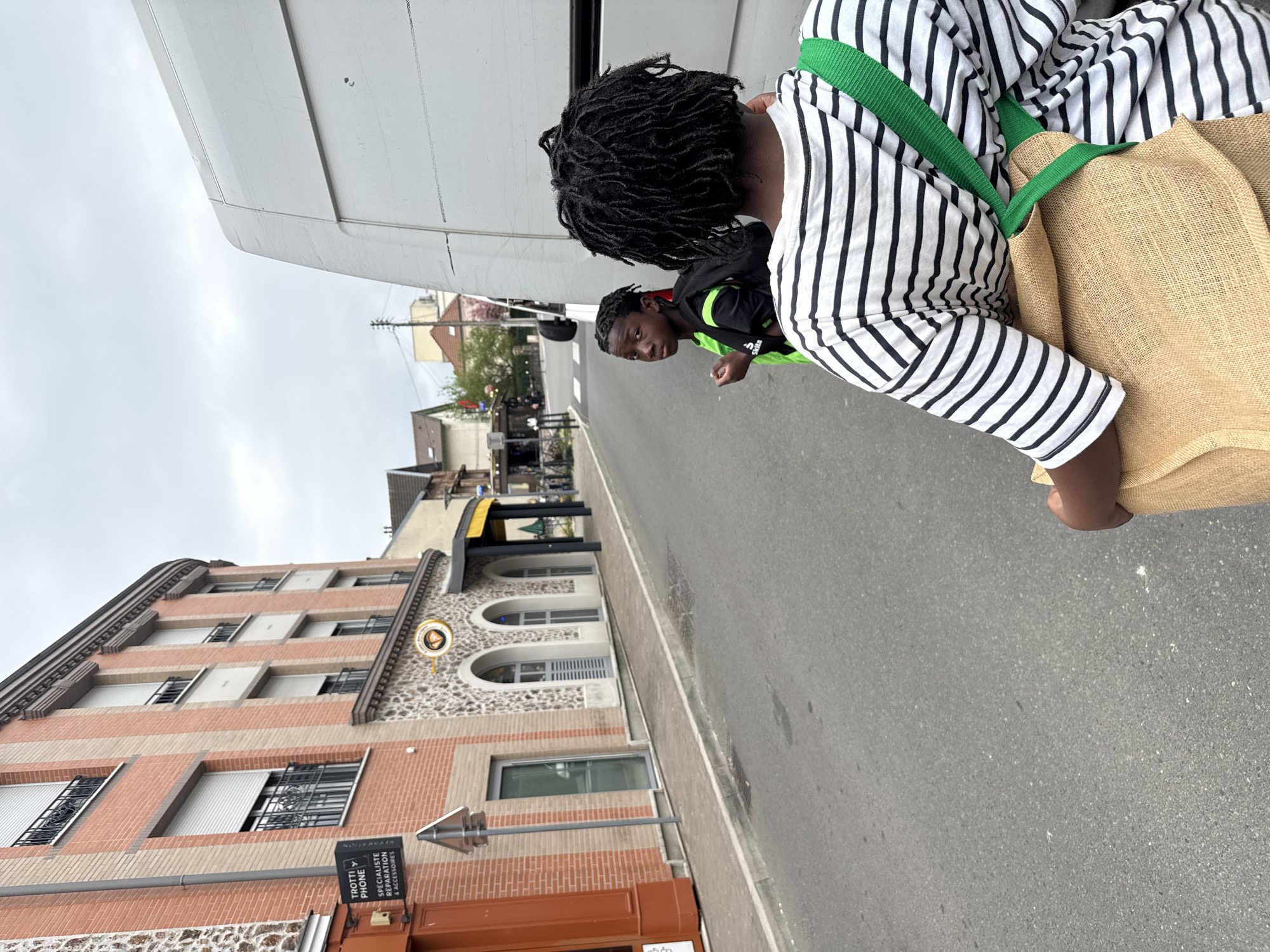 The image depicts a street scene in Domont, France. The foreground features two individuals: one with dreadlocks and wearing a black and white striped shirt with a green strap for a tan tote bag. The second person is a younger Black male, wearing a black jacket with green trim. They are standing on a grey asphalt road.
To the left of the road stands a multi-story building constructed of red brick and stone, with arched windows and metal railings. Signage is visible on the building, indicating "Trotti Phone" specializing in repair and accessories. In the background, other buildings and trees can be seen further down the street under an overcast sky. A large white vehicle appears in the top left corner.