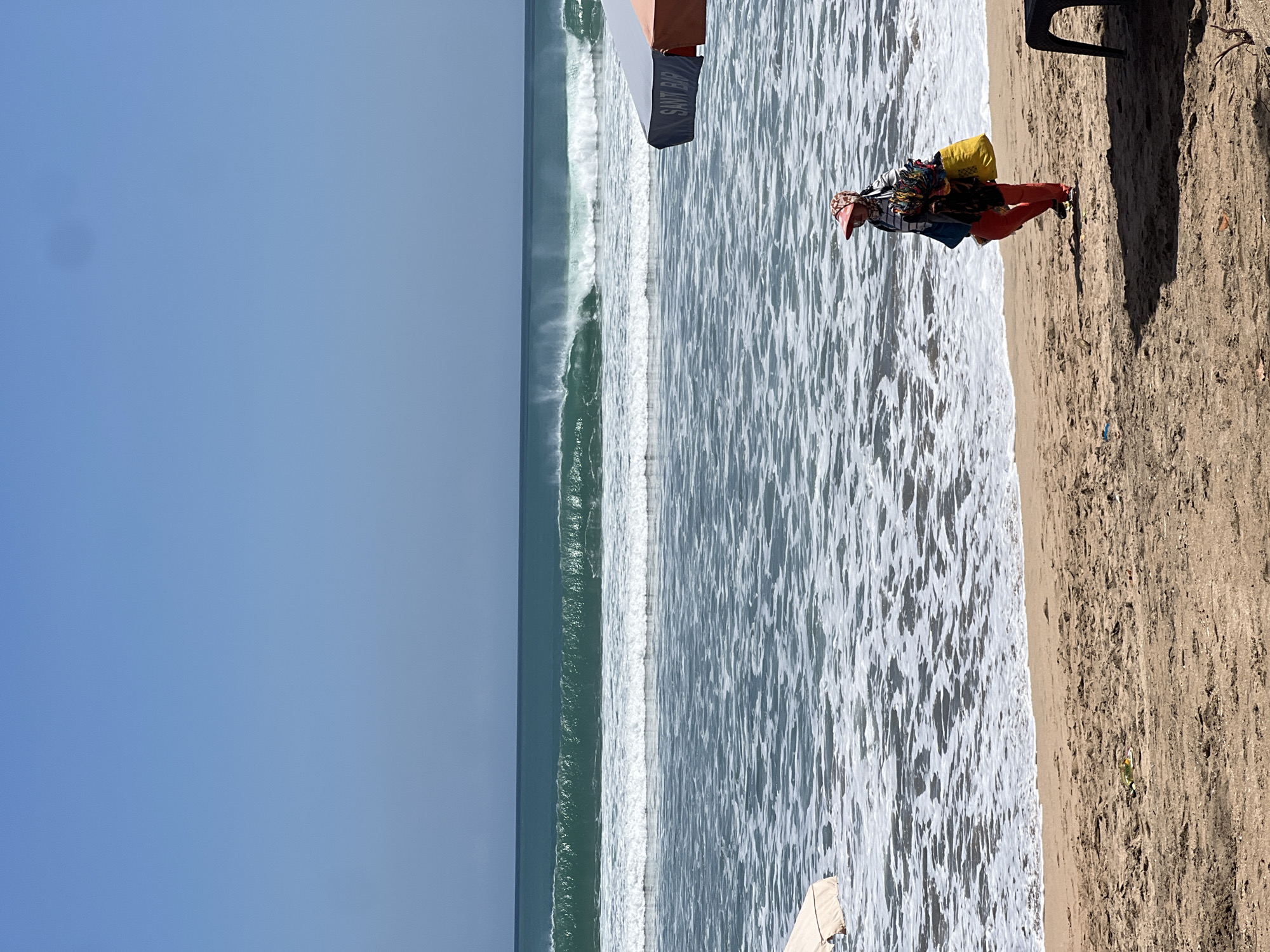 A woman is walking on a sandy beach in Kuta, Indonesia, with the ocean and waves in the background. The woman is wearing red pants, a striped shirt, and a pink sun hat. She is carrying a yellow bag over her shoulder and several colorful items are hanging from it. Two beach umbrellas are partially visible on the right side of the image, one with an orange and grey canopy and the other with a dark blue canopy with the text "SANT BIK" visible. The sky is clear and blue, and the water is a deep turquoise color with white waves.