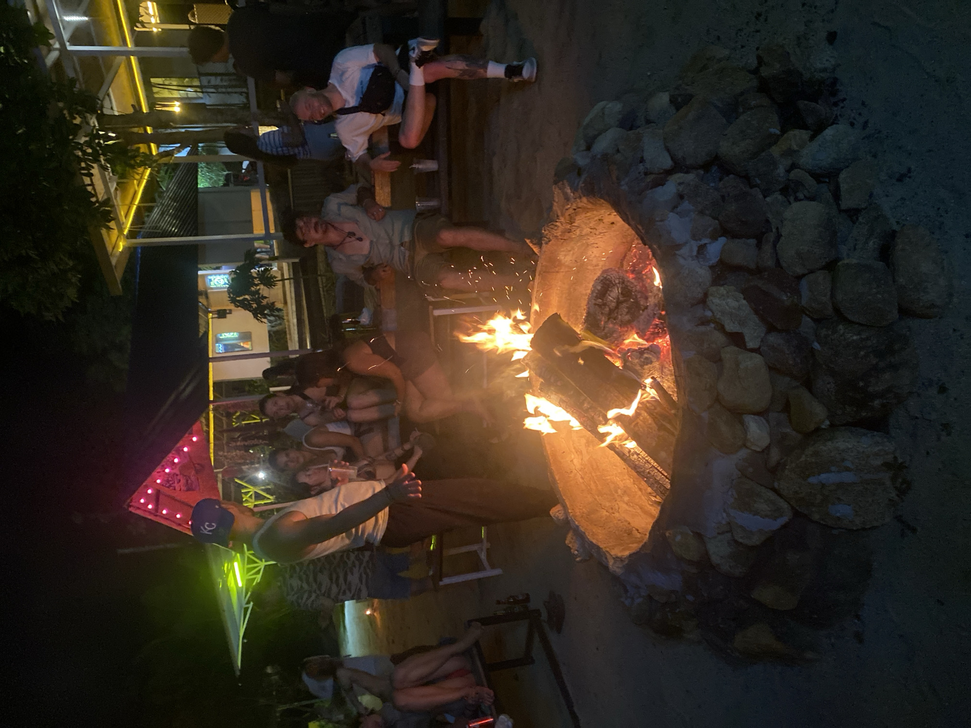The image shows a group of people gathered around a fire pit at night in Baan Hin Kong, Thailand. The fire pit is constructed of rocks and filled with burning logs, casting a warm glow. A group of people of diverse appearance and ages are sitting and standing near the fire, possibly engaging in conversation or socializing. A nearby structure with illuminated signage suggests a commercial establishment, perhaps a bar or restaurant, with some people sitting near the building. The overall environment appears to be casual and relaxed.