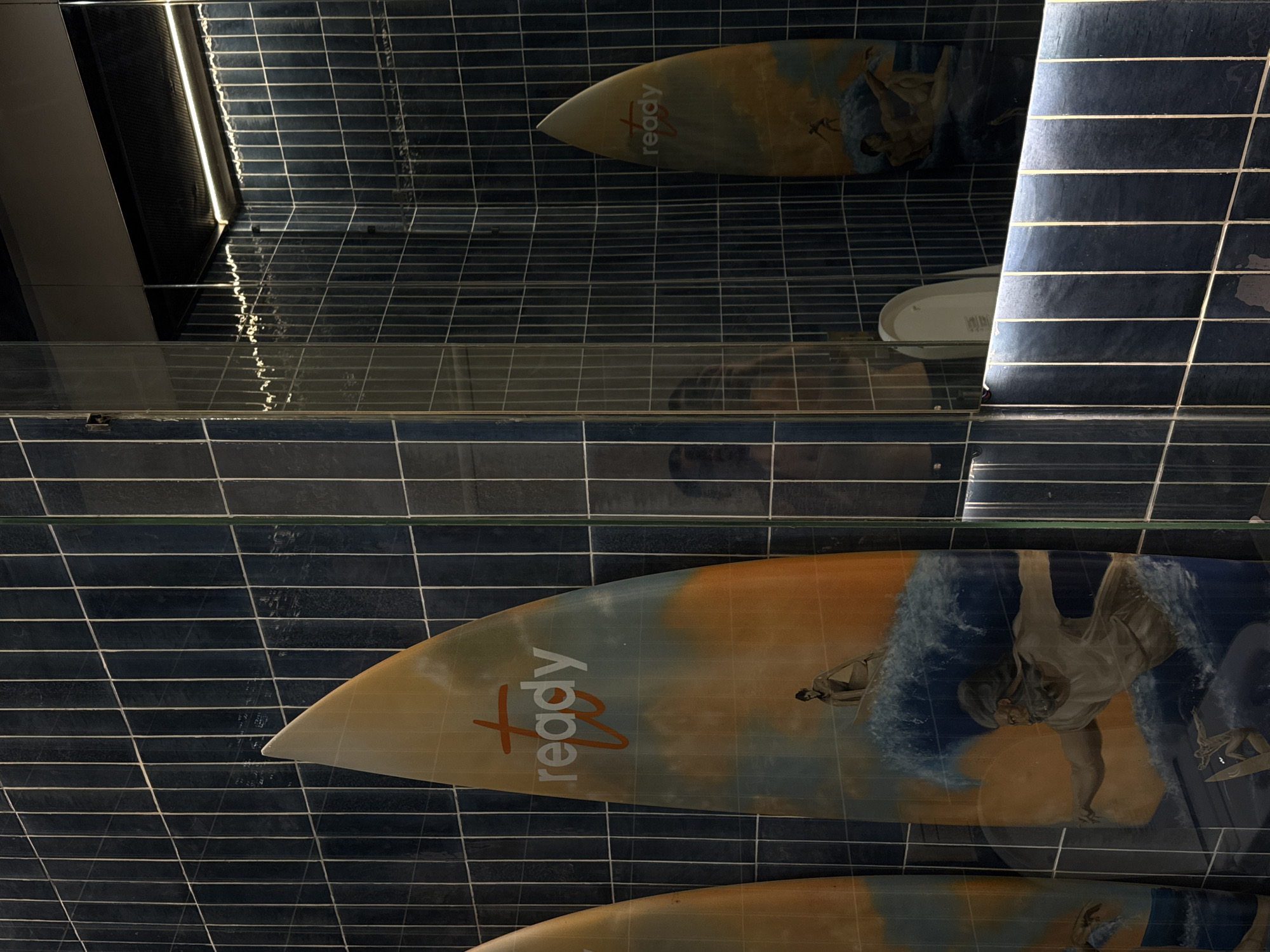 This image shows a bathroom interior with a dark blue tile wall in Kuta Selatan, Indonesia. The wall is decorated with surfboards painted with surfer imagery and the word "ready" in orange font. A mirror reflects the surfboards and part of the bathroom, including a toilet. The lighting appears to be artificial, and the overall perspective is low-angle, suggesting the photo was taken from a position near the floor.