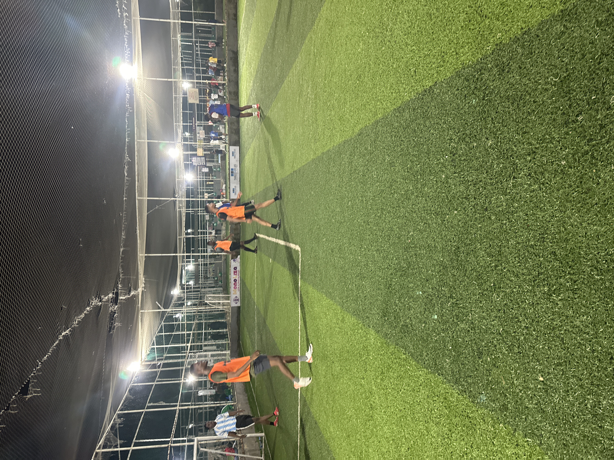 A group of men are playing soccer on an artificial turf field at night under artificial lights in the city of Abuja, Nigeria. The field is enclosed by fencing and netting, with bright lights illuminating the playing area. Several players are visible on the field, some in orange vests and shorts, others in blue and white striped jerseys. The men appear to be engaged in a casual game, with varying degrees of activity. The overall scene suggests a sports facility providing recreational opportunities in an urban setting.
