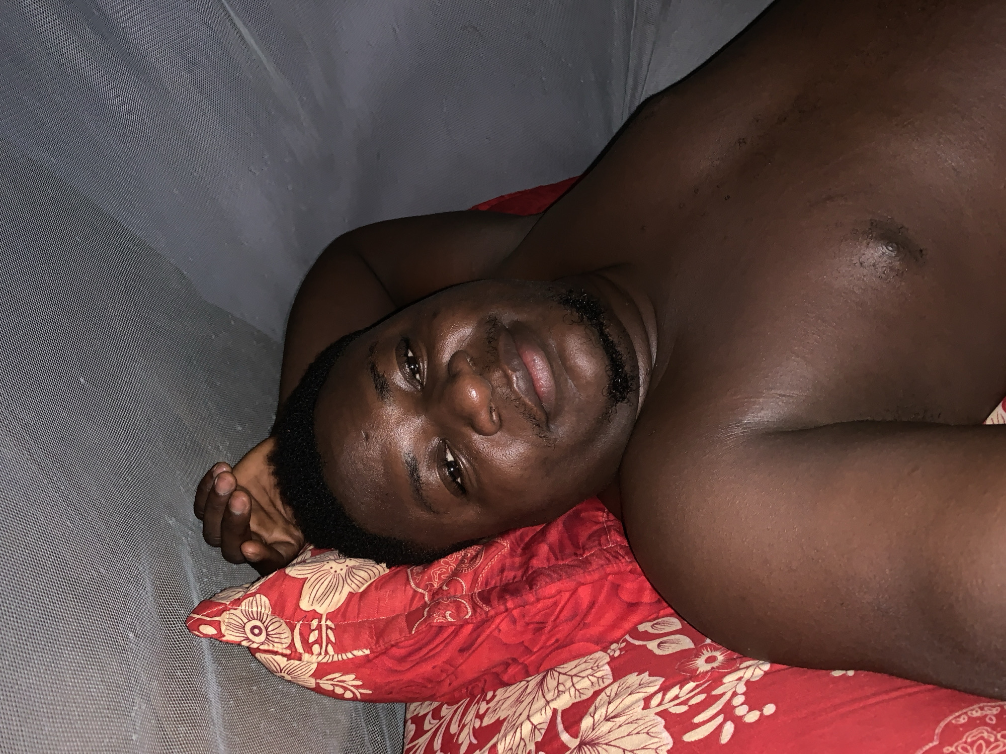 An adult male lies on his left side on a red floral-patterned pillow and bedspread. He is dark-skinned, with short black hair and a short beard. He is shirtless. The setting is indoors, possibly inside a mosquito net. It is impossible to verify if this image was taken in Akwanga, Nigeria.