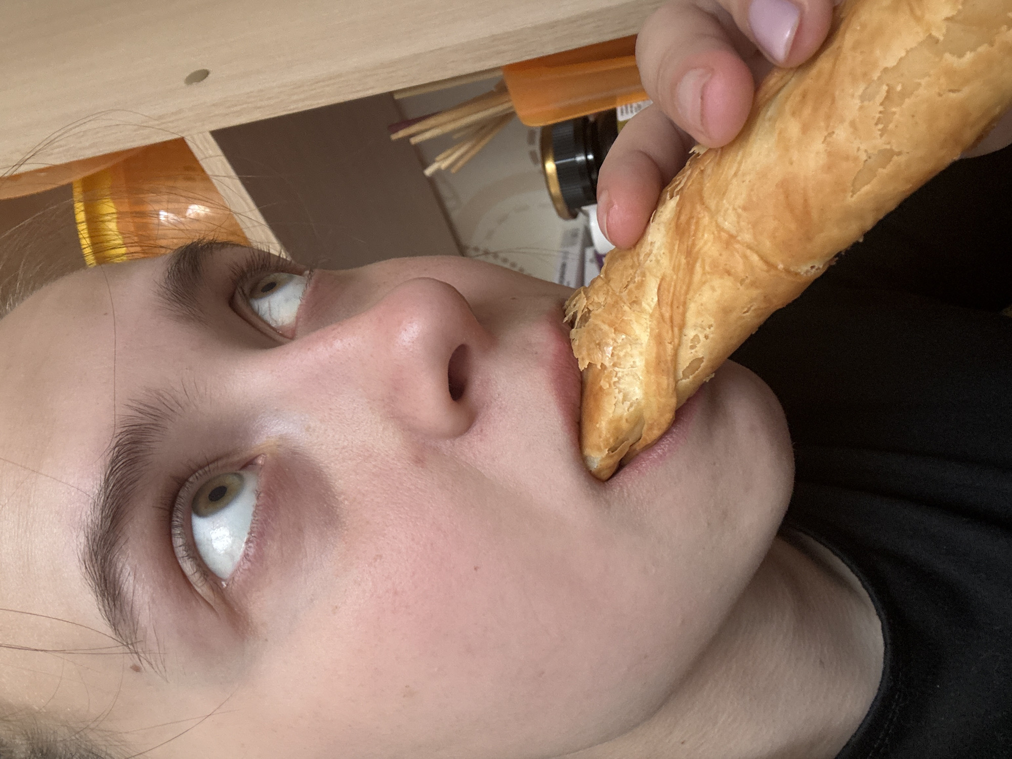 A close-up, upward-angled shot shows a person with fair skin, light brown eyebrows, and green eyes looking upwards, holding a pastry in their hand and placing it in their mouth. The pastry is golden brown and appears flaky. The person is wearing a black shirt.
Behind the person, under a wooden surface, are various objects including an orange container and cylindrical containers. The location or city of the photo cannot be determined from the provided image.