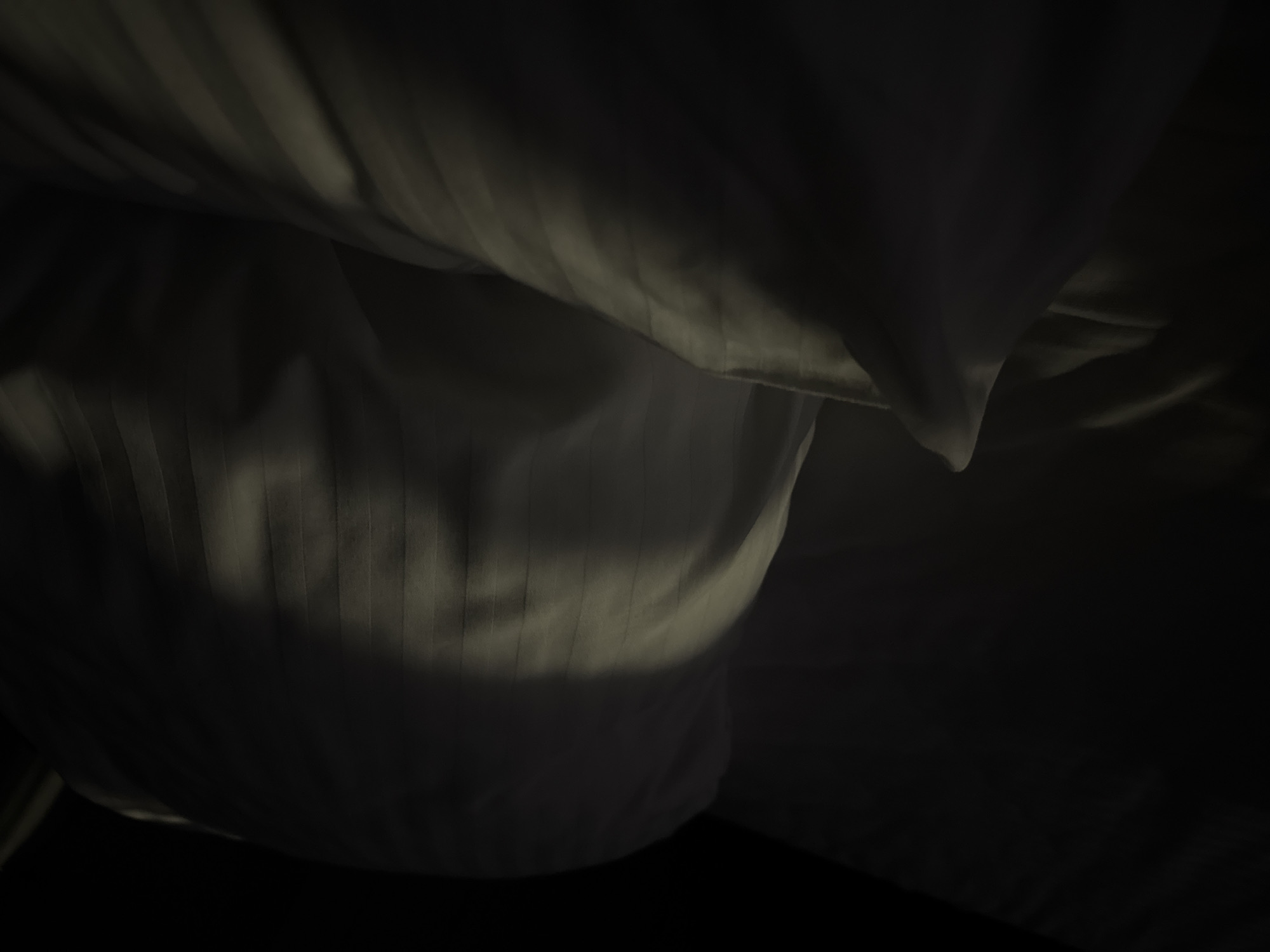 A close-up, low-angle shot captures a dimly lit scene of white striped bedding, likely pillows and sheets, within a room. A sliver of bright light cuts across the fabric from the top left, creating distinct shadows and highlights that emphasize the texture and folds of the material. The overall ambiance is dark and quiet, suggesting early morning or nighttime. The location is given as Doma 1096 Km, Russia. No people or specific actions are depicted.