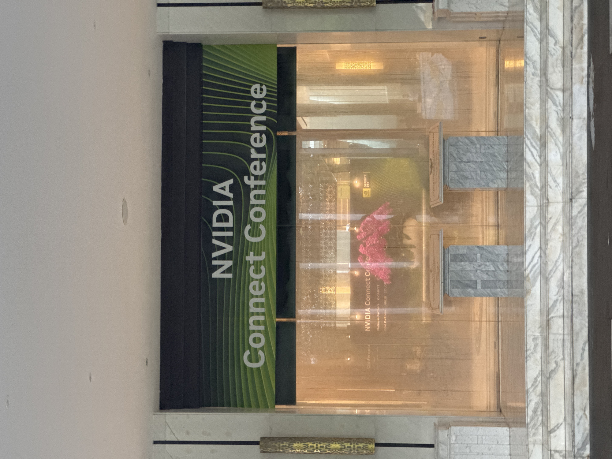 A vertically oriented image depicts the entrance to a venue, prominently featuring a large banner for the "NVIDIA Connect Conference." The banner has a dark background on the left and a vibrant green, striped pattern on the right, with "NVIDIA Connect Conference" written in white, uppercase letters.
Behind the banner are glass doors or windows, reflecting the interior of the building. Visible through and reflected on the glass are several details related to the conference. Another sign, partially obscured but showing "NVIDIA Connect Confer," indicates event sponsors and partners, specifically "Platinum Partners: GIGABYTE" and "Gold Partners ASUS COLOP." Also reflected is the "GEFORCE RTX" logo, likely from a display or sign inside. A large arrangement of pink flowers or a decorative plant is also reflected, adding a touch of color to the interior view.
The entrance itself is framed by elegant, light-colored marble pillars with distinct veining patterns. Between the pillars and the glass doors, metallic decorative panels with intricate patterns are visible. The overall setting appears modern and professional, indicative of a corporate event or exhibition space.
The image captures the entrance to an event, the NVIDIA Connect Conference, held in a building featuring contemporary design elements like glass and marble. The location context provided places this scene in Kuta Selatan, Indonesia. The time of day is not explicitly clear, but the interior lighting suggests it could be during operational hours.