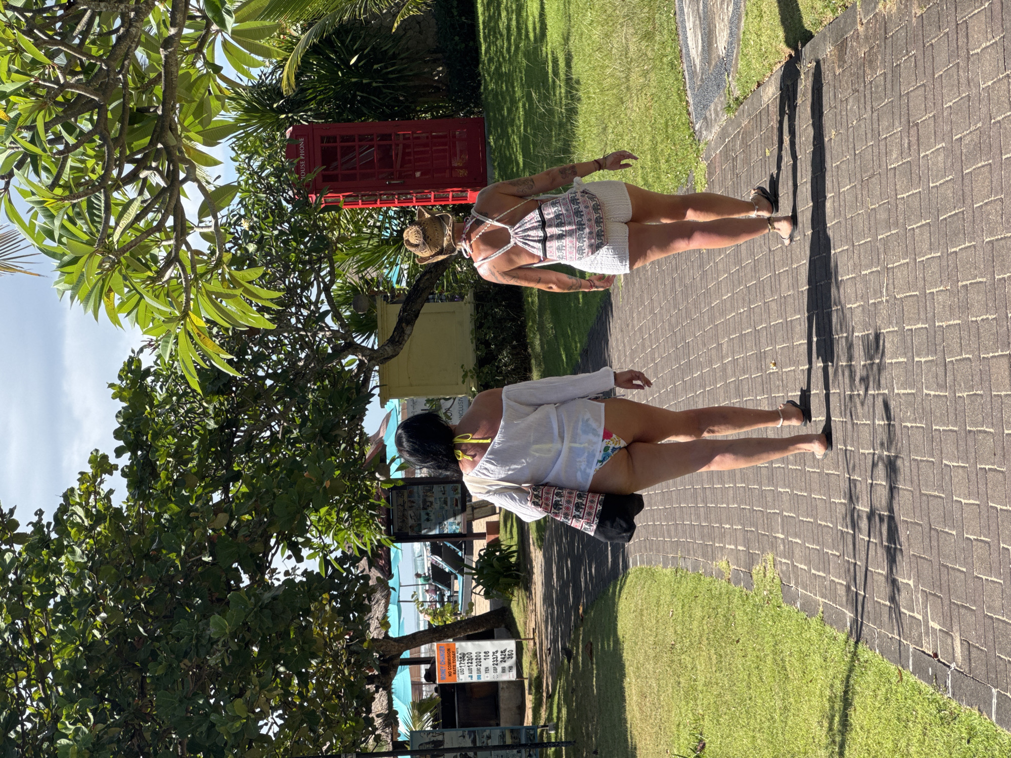 Two women walk along a brick paved path in Kuta Selatan, Indonesia. The woman in the foreground wears a white long-sleeve shirt, bikini bottoms, sandals and carries a patterned bag on her shoulder. The woman in the background wears a white top with shorts, sandals, and a hat. A red phone booth is visible in the background along with green grass, trees, a sign detailing exchange rates. The setting is outdoors during daylight hours.