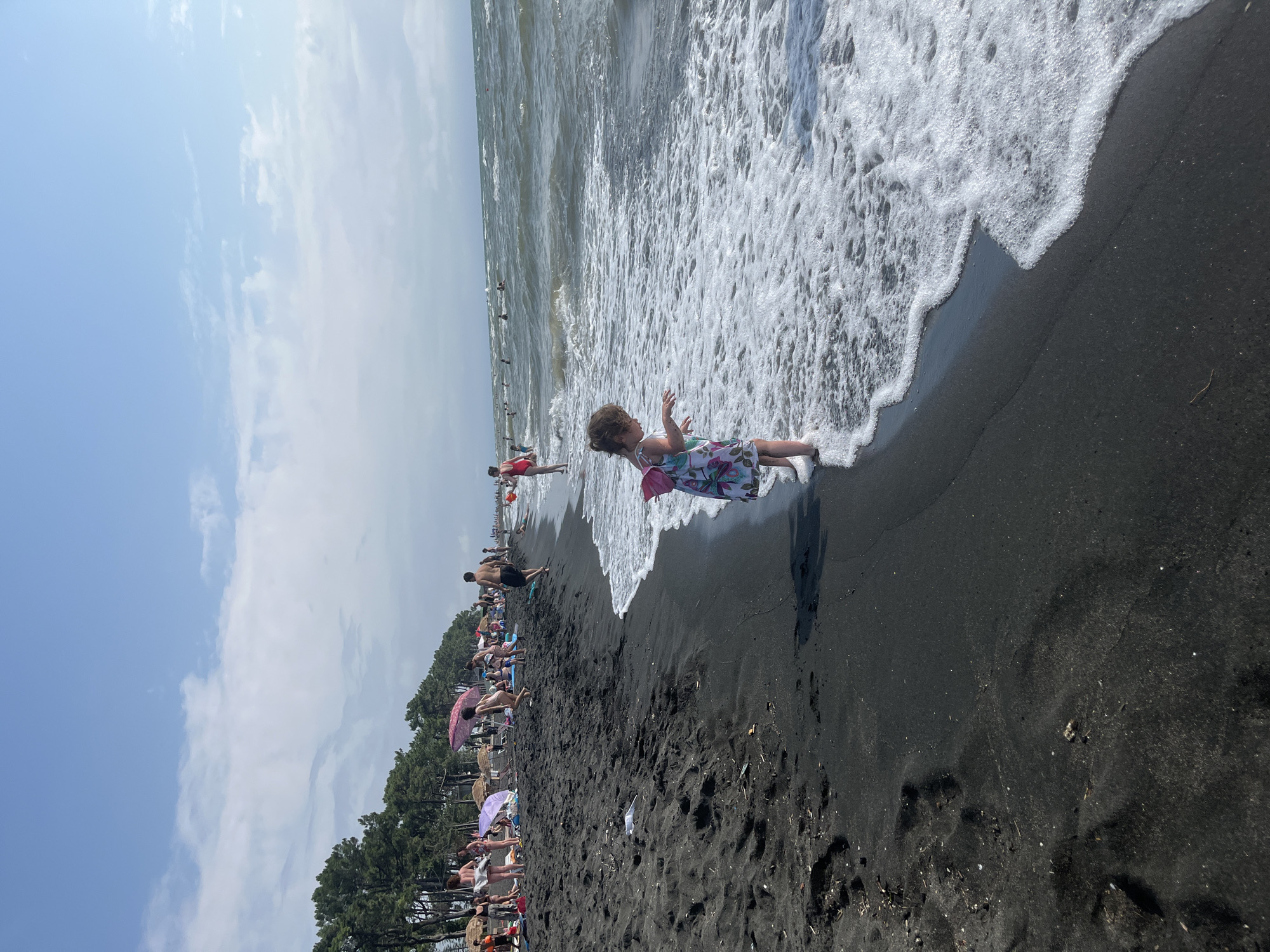 The image depicts a sunny day at Ureki Beach in Georgia, known for its black sand. The foreground features a young girl in a floral dress and a pink backpack, standing at the edge of the water where waves are gently washing onto the shore. The wet black sand is visible around her feet, with some footprints and debris scattered on the dry sand further back.
In the midground, the beach is populated with numerous people, some lounging on towels, others walking or playing. Beach umbrellas, both colorful and straw, dot the shoreline, providing shade. A line of evergreen trees forms a backdrop behind the beachgoers.
The background shows a hazy sea under a bright sky with scattered white clouds. The water is choppy, indicating a moderate breeze. More people are visible in the water, swimming or wading. The overall scene captures a lively summer day at a popular beach destination.