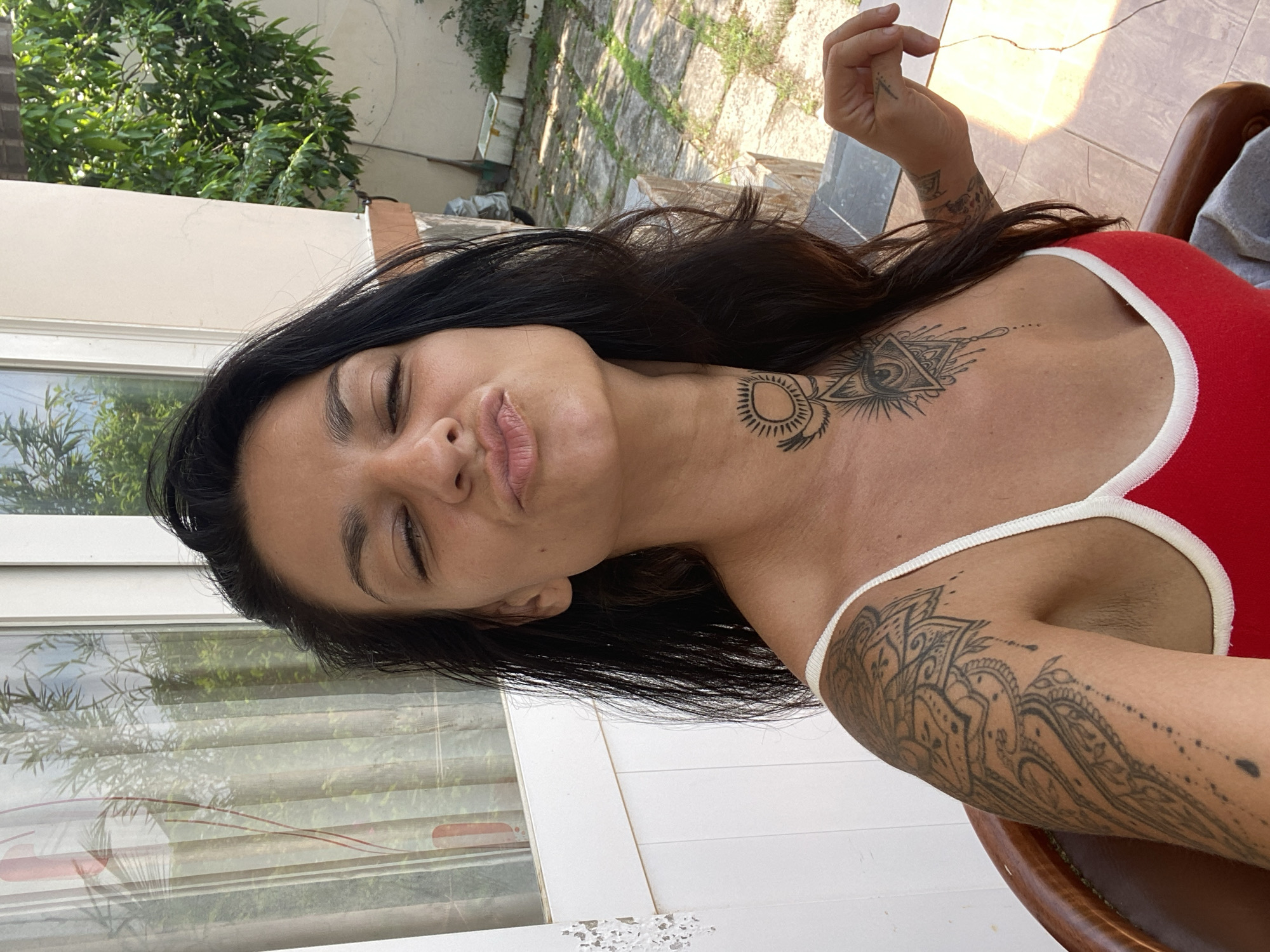 A woman with long, dark hair is posing for a selfie outdoors. She is wearing a red tank top with white trim. She has tattoos on her left arm, chest, and neck. Her eyes are closed and her lips are pursed. The background shows a white wall, a window with curtains, and some greenery. The flooring appears to be stone tiles. The scene is likely in Mũi Né, Vietnam, given the vegetation and architecture commonly found in the region.