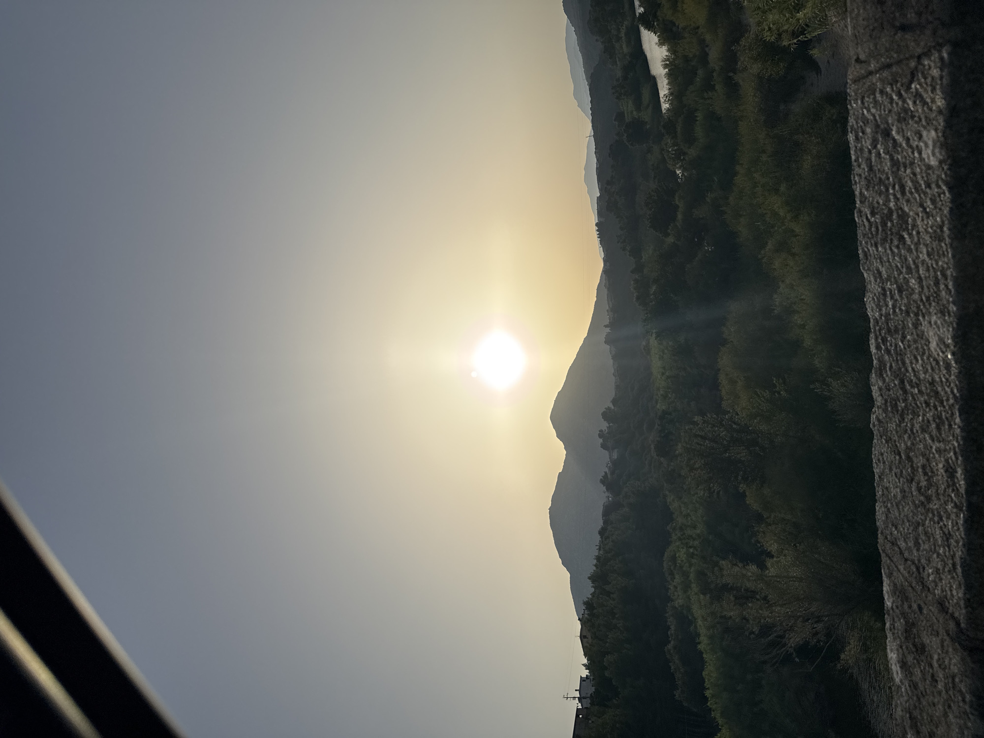 The image depicts a serene natural landscape under a bright, hazy sun, likely during sunrise or sunset. The sun, a radiant orb, is positioned in the upper center of the frame, casting a soft, diffused light across the scene. In the foreground, a rough, textured stone surface occupies the bottom edge, suggesting a viewpoint from a bridge or wall. Beyond this, rolling hills covered in lush, dark green vegetation dominate the mid-ground. A winding river or body of water is visible on the right side, reflecting the ambient light. Silhouettes of distant mountains are visible on the horizon, adding depth to the composition. A few scattered structures, appearing to be houses, are present on the left side, nestled amongst the trees, hinting at a nearby settlement. The overall atmosphere is tranquil and picturesque, characteristic of a Mediterranean environment, possibly near the city of Orosei, Italy, known for its scenic natural beauty.