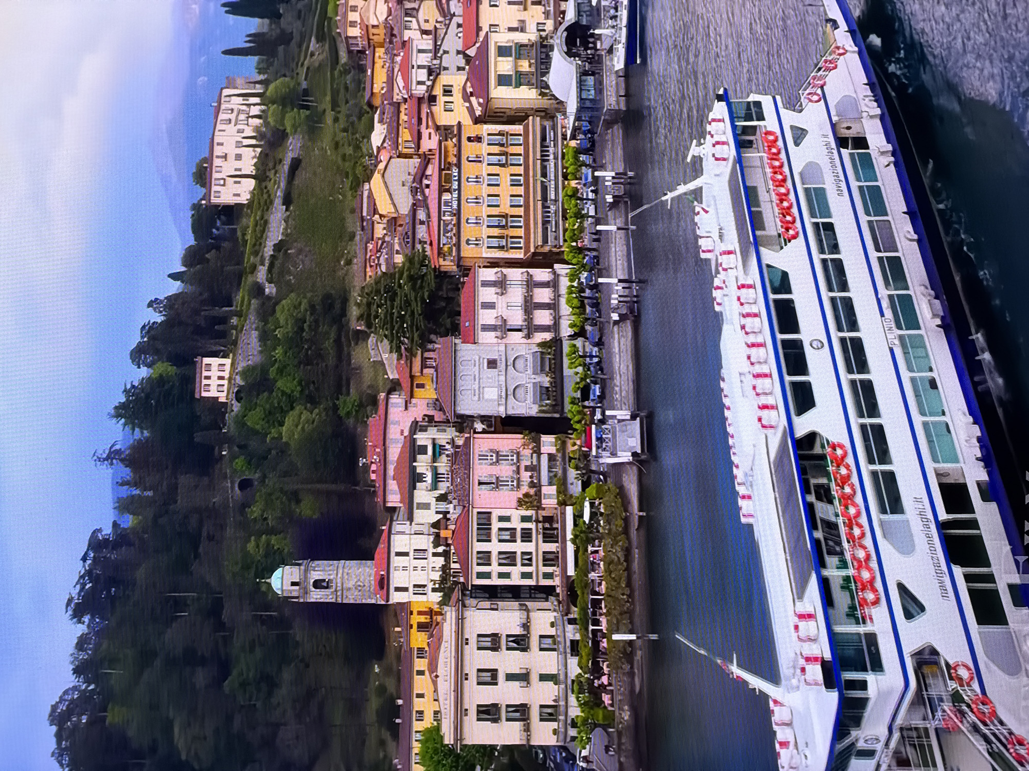 The image shows a panoramic view of a scenic waterfront town nestled against a lush green hillside. In the foreground, a large white ferry with blue trim and multiple decks is docked in the water. The ferry displays the text "navigazionelaghi.it" and "PLINIO" on its hull. Life preservers in red and white are visible along its sides.
Behind the ferry, a waterfront promenade is lined with a row of colorful buildings. These buildings exhibit a variety of architectural styles, with predominantly pastel facades in shades of yellow, pink, and cream, topped with red-tiled roofs. Many of these buildings appear to be hotels or businesses, with signs visible, such as "HOTEL FLORENCE" and "HOTEL DU LAC." Outdoor seating areas with tables and chairs are set up along the waterfront in front of some establishments.
Further up the hillside, more buildings are visible, interspersed with trees and greenery. A prominent church tower with a distinctive green dome stands out against the backdrop of the hill. Higher up, larger villas or mansions with pale facades are situated on the elevated terrain. The hillside is densely covered with trees, creating a rich green landscape that slopes towards the water. The sky is a pale blue with some wispy clouds, and distant mountains can be seen in the background, veiled in a light haze.
The scene is devoid of any specific identifiable human figures in action, but hints of human presence are suggested by the outdoor seating and the functioning ferry. The overall atmosphere is serene and picturesque, characteristic of a European lakeside resort town.
The provided location of Chirchik, Uzbekistan, does not align with the visual cues in the image, which strongly suggest a European setting, likely on a lake or large body of water, with architecture common to Italian lake towns.