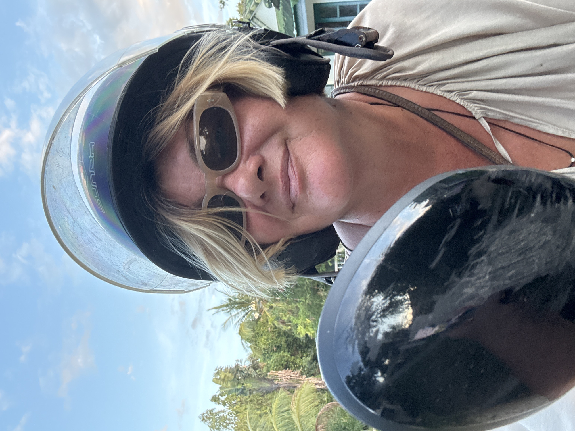A medium shot, taken from a low angle, depicts a woman wearing a black helmet and light-colored sunglasses. The helmet has a clear face shield pulled down. She is wearing a tan-colored top, and a necklace is visible. The background includes a blue sky with some clouds, green trees, and a building with blue shutters. The image appears to be taken from the perspective of someone on a motorcycle, with part of the motorcycle's mirror visible in the foreground. The picture may have been taken in Baan Bo Phut, Thailand, based on the possible location information.