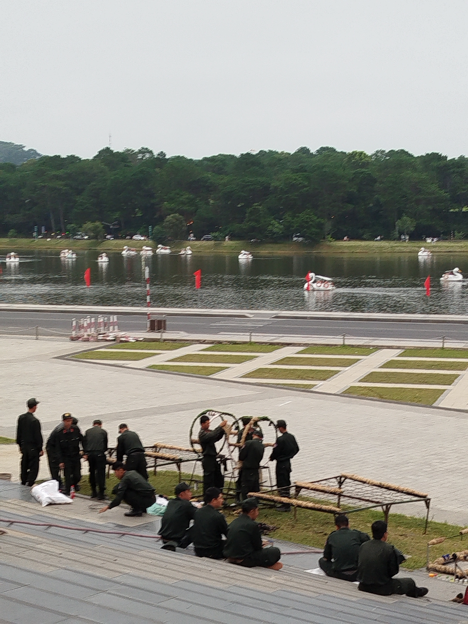 The image captures a lakeside scene in Da Lat, Vietnam. In the foreground, a group of people, some in dark uniforms and others in civilian clothing, are gathered on tiered steps. Some are seated, while others stand or crouch, appearing to be engaged in a collective activity, possibly preparing for or dismantling an event. One individual in uniform is crouched, pointing towards the ground, while others are observing or working with what appear to be decorative elements or equipment.
Further back, beyond a paved area and a road, a lake stretches out. Several swan-shaped pedal boats are visible on the water, indicating a recreational setting. Red flags are planted in the lake and on the shore, adding a splash of color to the scene. A dense line of evergreen trees borders the far side of the lake, creating a natural backdrop. The sky is overcast, suggesting a cloudy day. The overall impression is of a public space being utilized for an event or a gathering, with a blend of organized activity and leisure.