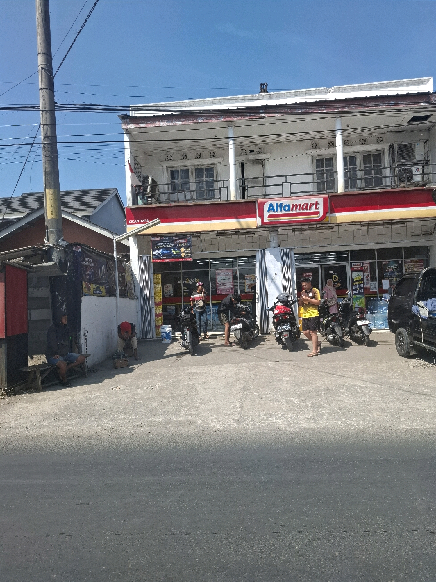The image captures a street scene in Selajambe, Indonesia, featuring a two-story building with a prominent Alfamart convenience store on the ground floor. The building's facade is white with red and yellow trim. The Alfamart signage is clearly visible. In front of the store, several motorcycles are parked.
On the left side of the image, a smaller, makeshift structure with a corrugated metal roof is present. Two individuals are seated on a wooden bench outside this structure, one appearing to be looking away and the other facing forward. A utility pole with numerous electrical wires is positioned to the left of the building.
In the foreground, a paved road with some debris is visible. The sky is clear and blue. A black car is parked to the right of the Alfamart. People are seen standing and interacting near the motorcycles in front of the store. The overall environment suggests a commercial area with residential elements nearby.