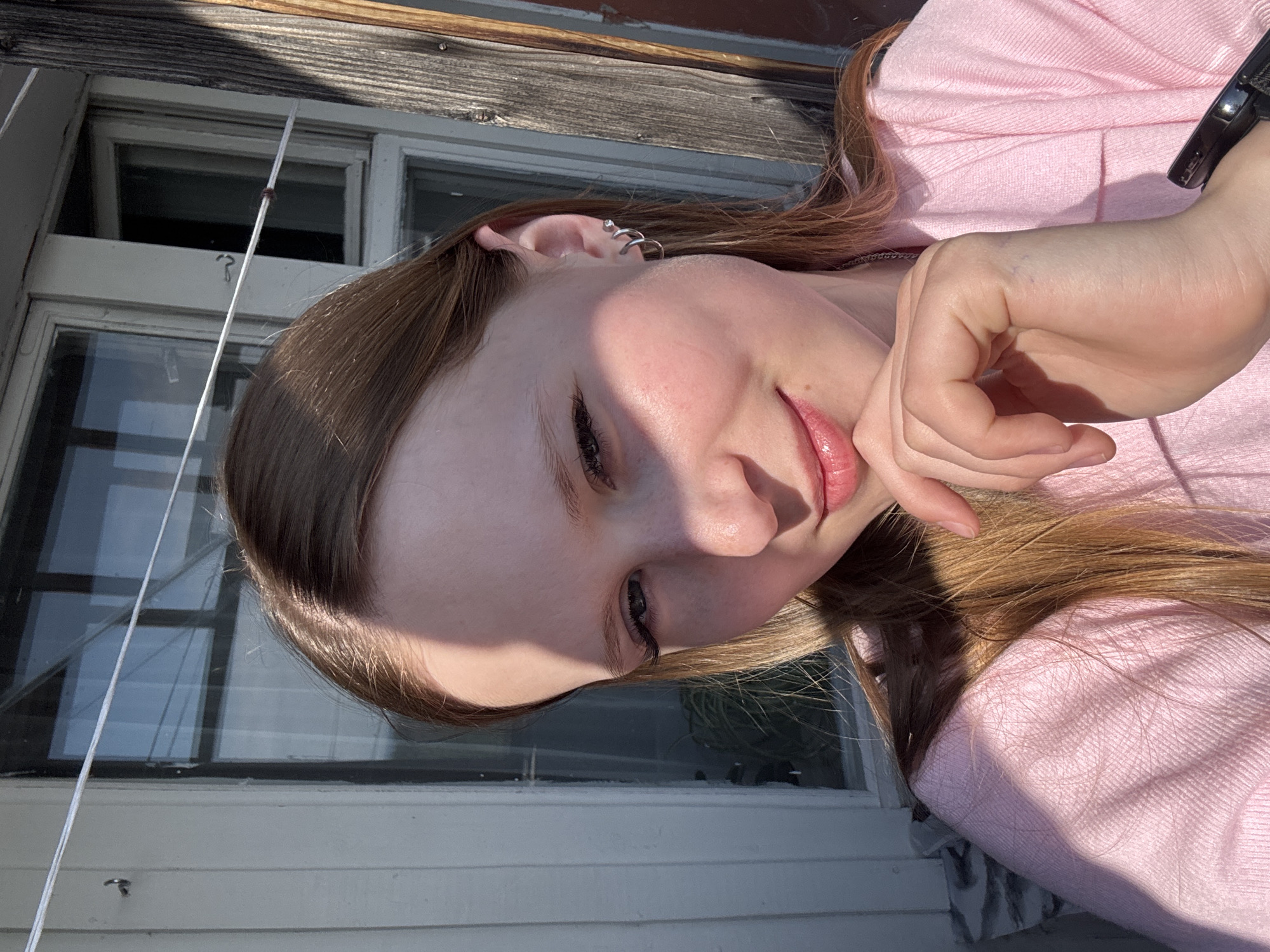 A close-up, tilted portrait captures a young woman with long, light brown hair and a fair complexion. She is wearing a light pink long-sleeved top and has an intricate silver helix earring on her left ear. She looks down with a gentle, soft smile, her lips slightly parted, and her right hand rests near her chin.
The background reveals details of an old building, possibly in Chaikovsky, Russia. To her left, white-framed windows with dark panes are visible, some appearing slightly ajar. Above the windows, there is a weathered wooden beam. Below the windows, white horizontal siding can be seen. A thin white string or cord hangs vertically near the window frame, with a small, dark object tied to it, possibly a hook or a weight.
The scene is illuminated by strong, direct sunlight, casting harsh shadows across the woman's face, hair, and the building, suggesting it is midday or early afternoon on a clear, sunny day. The woman's expression conveys a sense of calm and contentment. There is no visible text in the image.