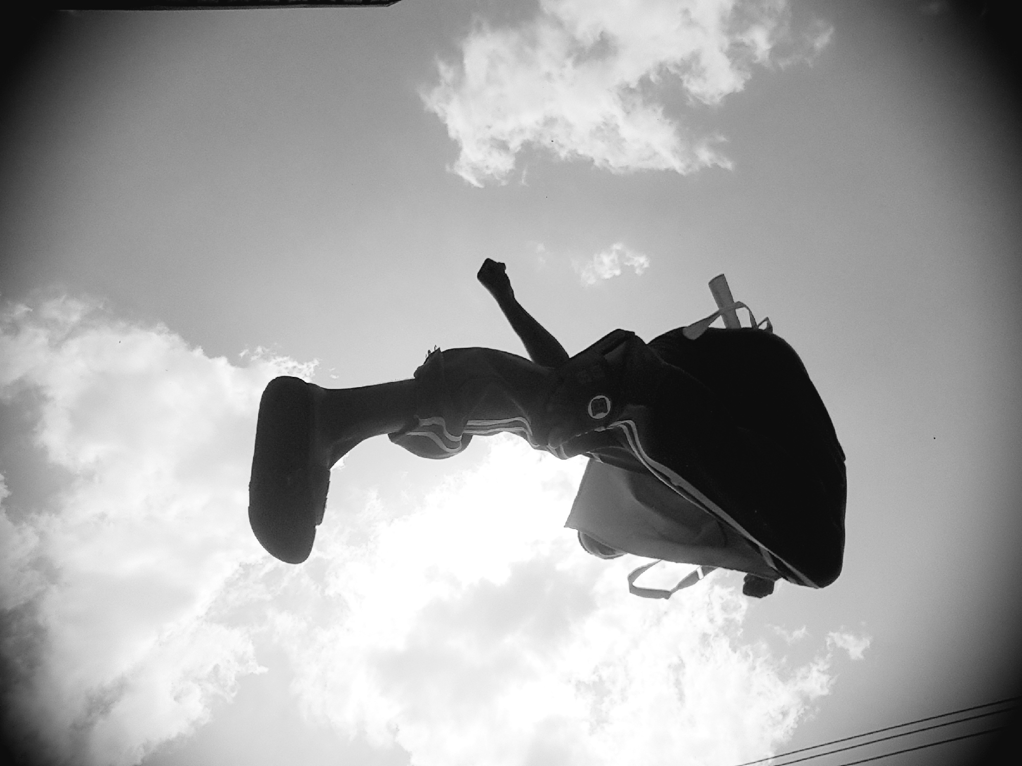 A low-angle, black and white photograph captures a person mid-air, their body inverted against a cloudy sky. The subject appears to be wearing athletic clothing with striped accents on the sleeves and legs, and a backpack is slung over their shoulders. One leg is extended upwards, and the other is bent. The background consists of a bright, overcast sky with scattered clouds. In the lower left corner, faint parallel lines suggest utility wires. The overall impression is of a dynamic action shot, possibly a jump or a gymnastic maneuver, with the city of Narayi, Nigeria, implied as the location.