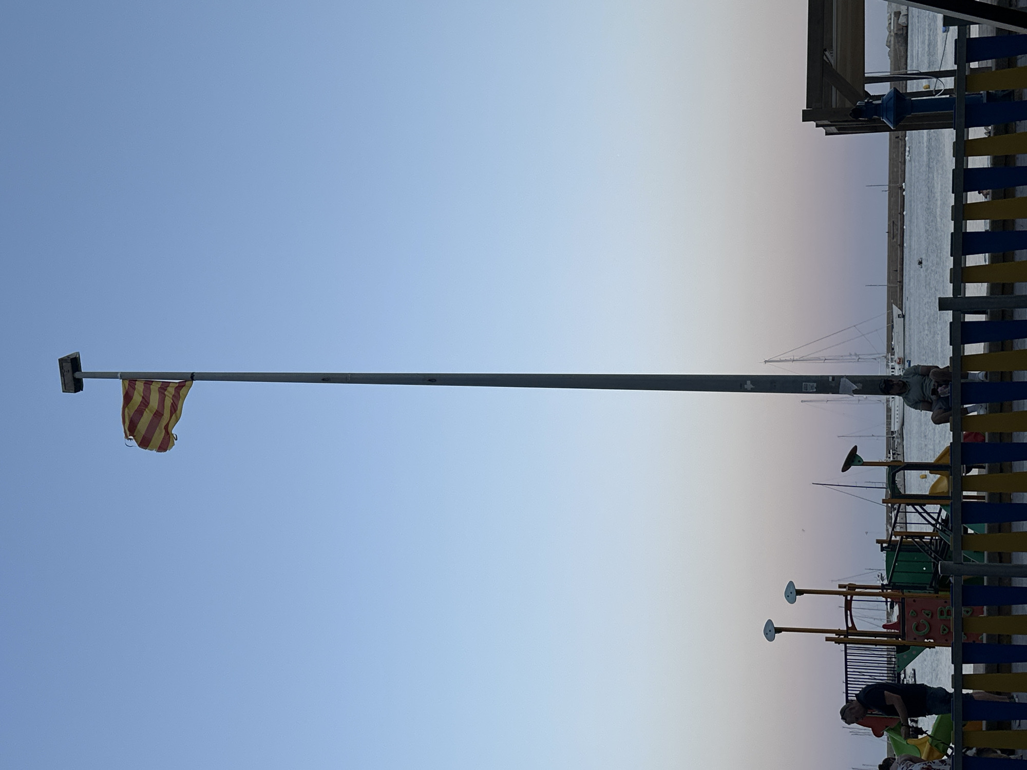 The image depicts a coastal scene with a prominent flagpole flying a striped flag, likely the Catalan flag, against a gradient sky transitioning from blue to pale pink. In the foreground, there is a children's playground with colorful structures and a blue, yellow, and red fence. To the left, a man wearing a dark shirt and blue jeans is interacting with the playground equipment. Further back, several sailboats are anchored in the water, with a breakwater visible in the distance. On the right side of the image, a covered seating area with blue lighting is present. The overall setting suggests a public space, possibly a waterfront promenade or park in Sant Feliu de Guíxols, Spain, during the evening or early morning.