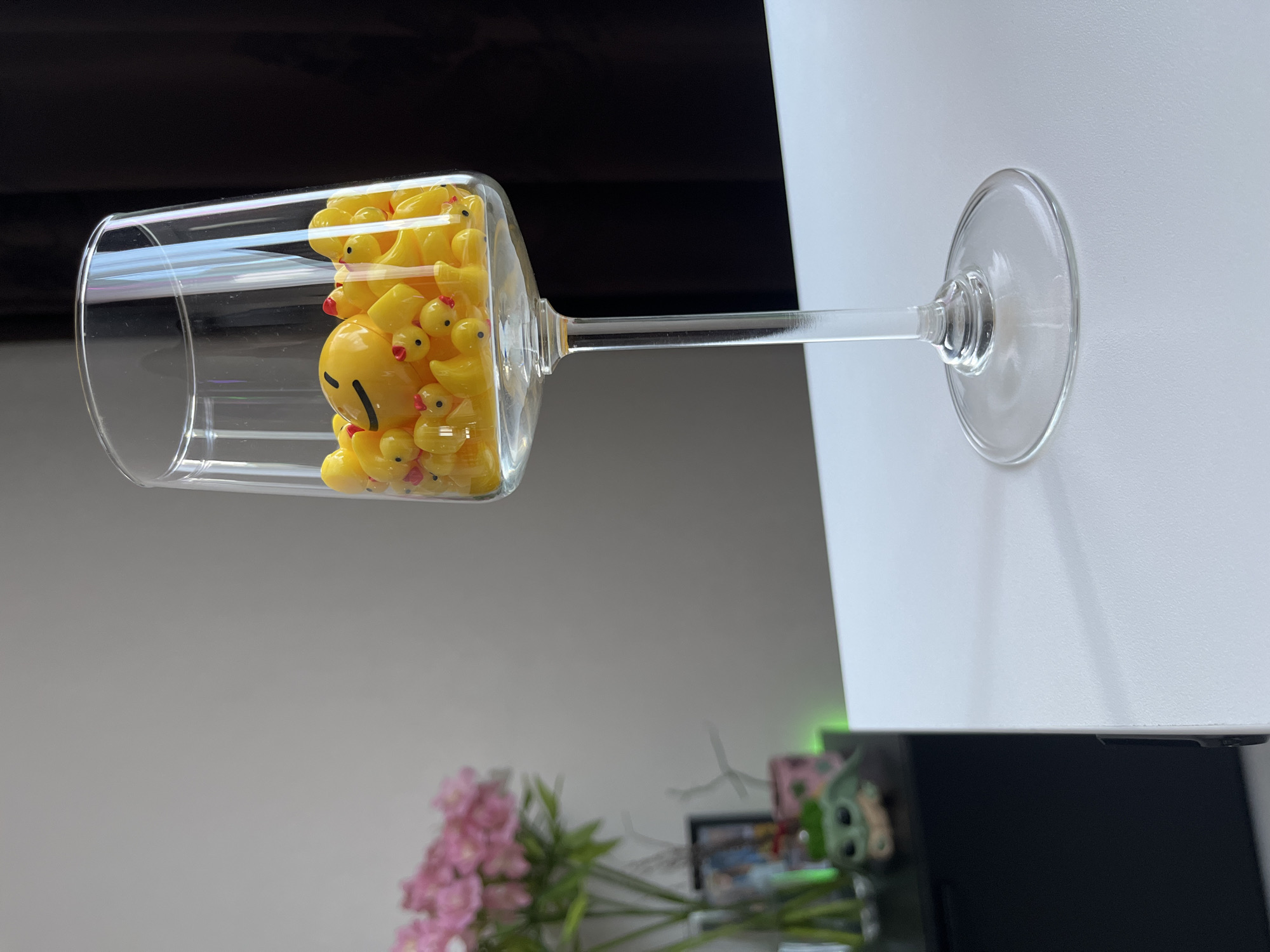 A tall, clear champagne flute is filled with numerous small yellow rubber ducks and a single larger yellow duck with a smiley face drawn on it. The flute rests on a white surface, likely a table. In the blurred background to the left, a vase of pink flowers is visible on a dark surface, beside a small figure with green accents. A dark curtain is behind the flute on the right. The image does not contain information about the city of Babruysk, Belarus.