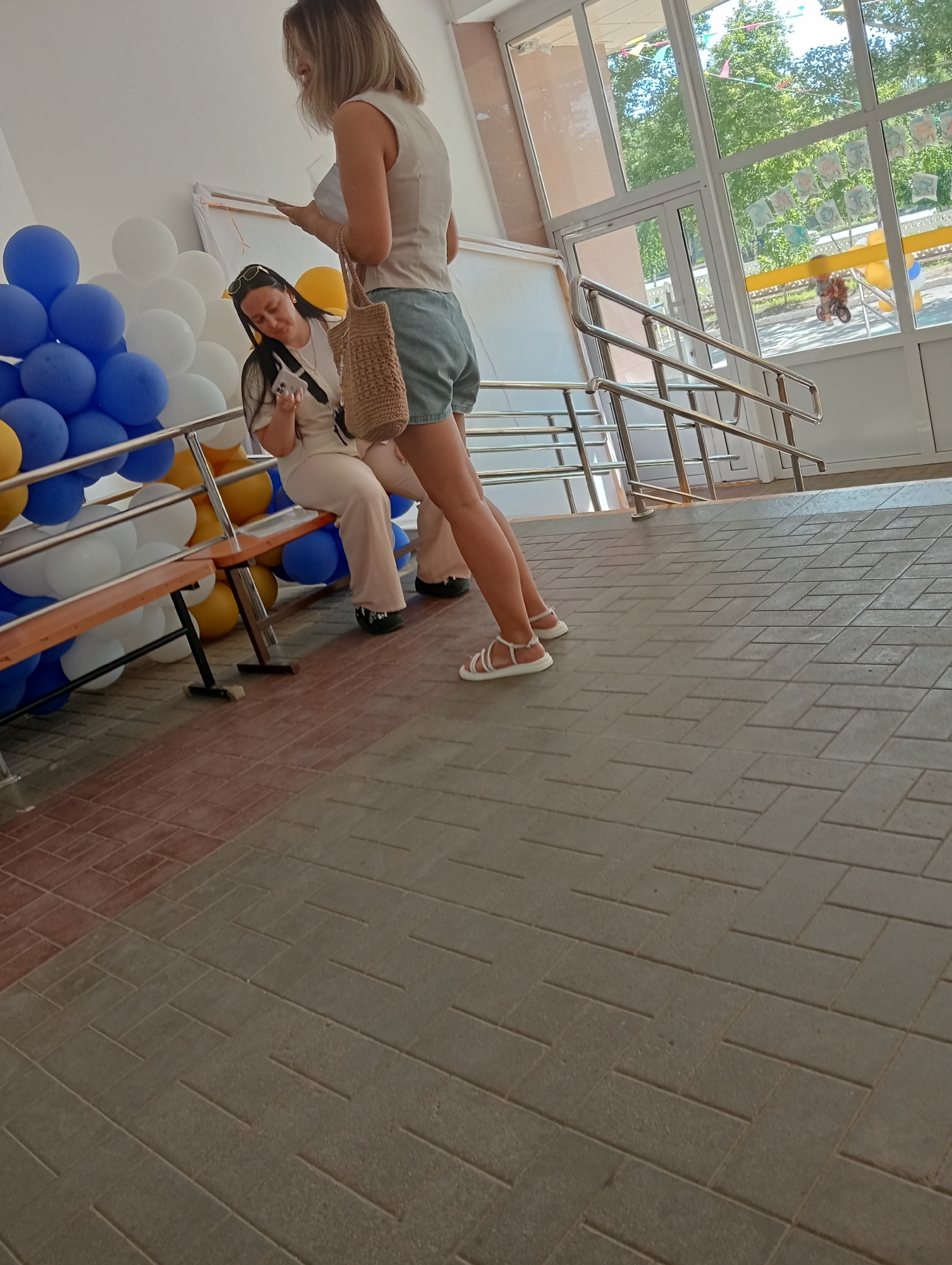Two women are near a ramp with a metal railing. One woman is standing and holding a paper and a bag. The other woman is sitting on a wooden bench, looking at her phone. Balloons are on the left side of the image. The ground is paved with gray and red tiles. The background shows a building with large windows and trees outside.