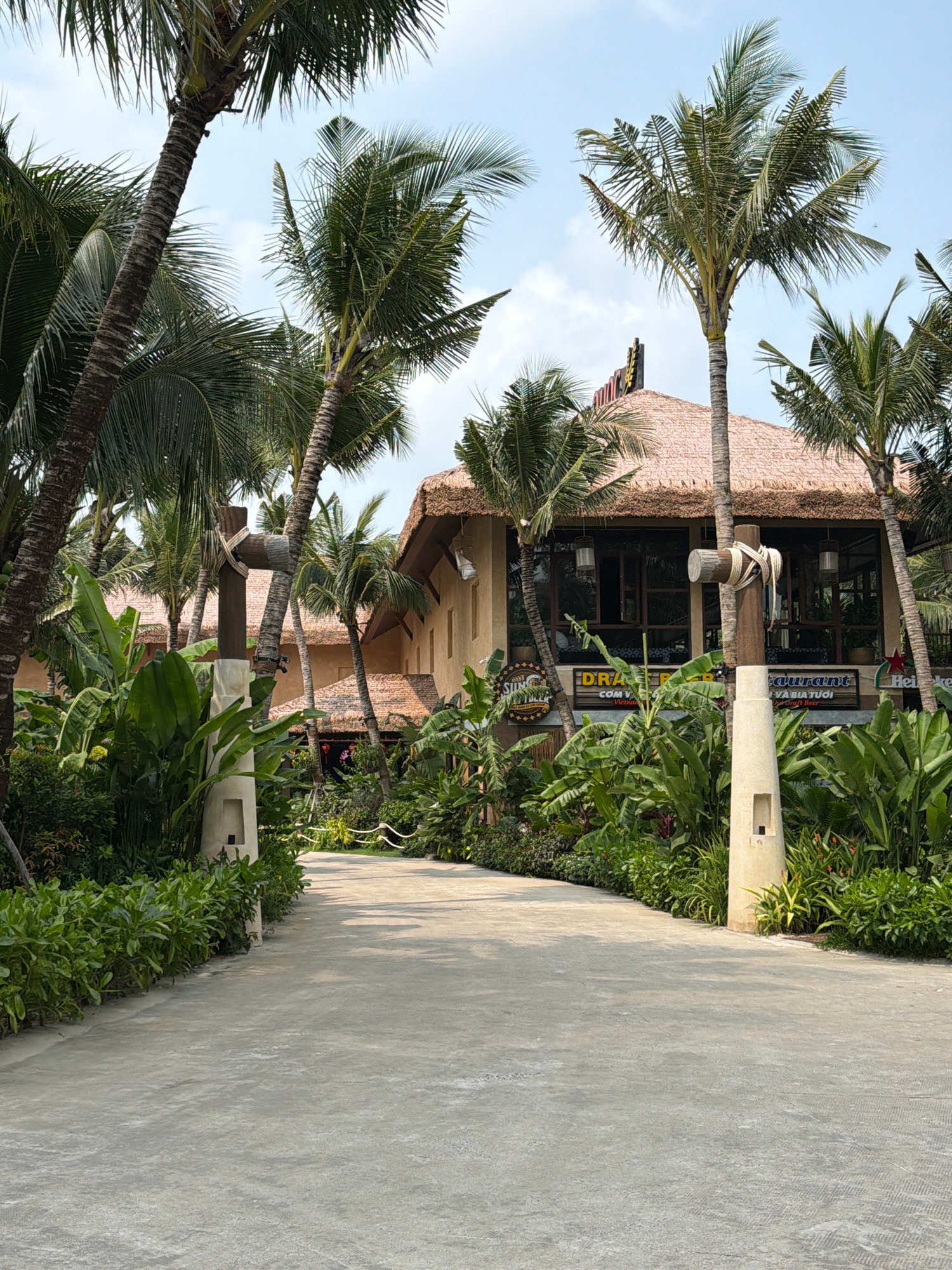 A sun-drenched pathway leads towards a thatched-roof building, likely a restaurant, in Phú Quốc, Vietnam. Lush tropical foliage, including palm trees and large-leafed plants, flanks the path and surrounds the building. The building has large windows and a sign that reads "DRA-GER RESTAURANT" and "CƠM VIỆT NAM". A wooden post with rope detailing stands on the left side of the path. The sky is a light blue with scattered clouds. The overall scene evokes a relaxed, tropical atmosphere.