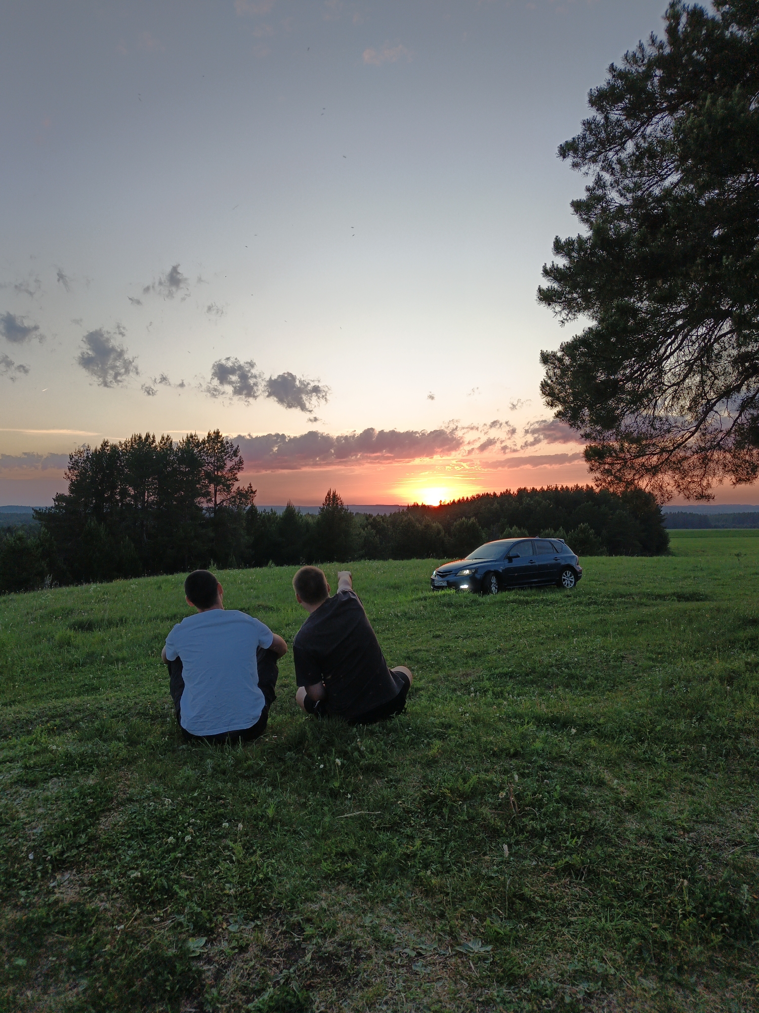 Two people are sitting on a grassy hillside facing a sunset. The person on the left is wearing a light-colored shirt, and the person on the right is wearing a dark shirt and pointing towards the sunset. Behind them, there is a dark-colored car parked in the grass. A line of trees is visible on the horizon, and a large tree is on the right side of the frame. The sky is a mix of light blue, pink, and orange. The location is most likely near the city of Чумна, Russia.