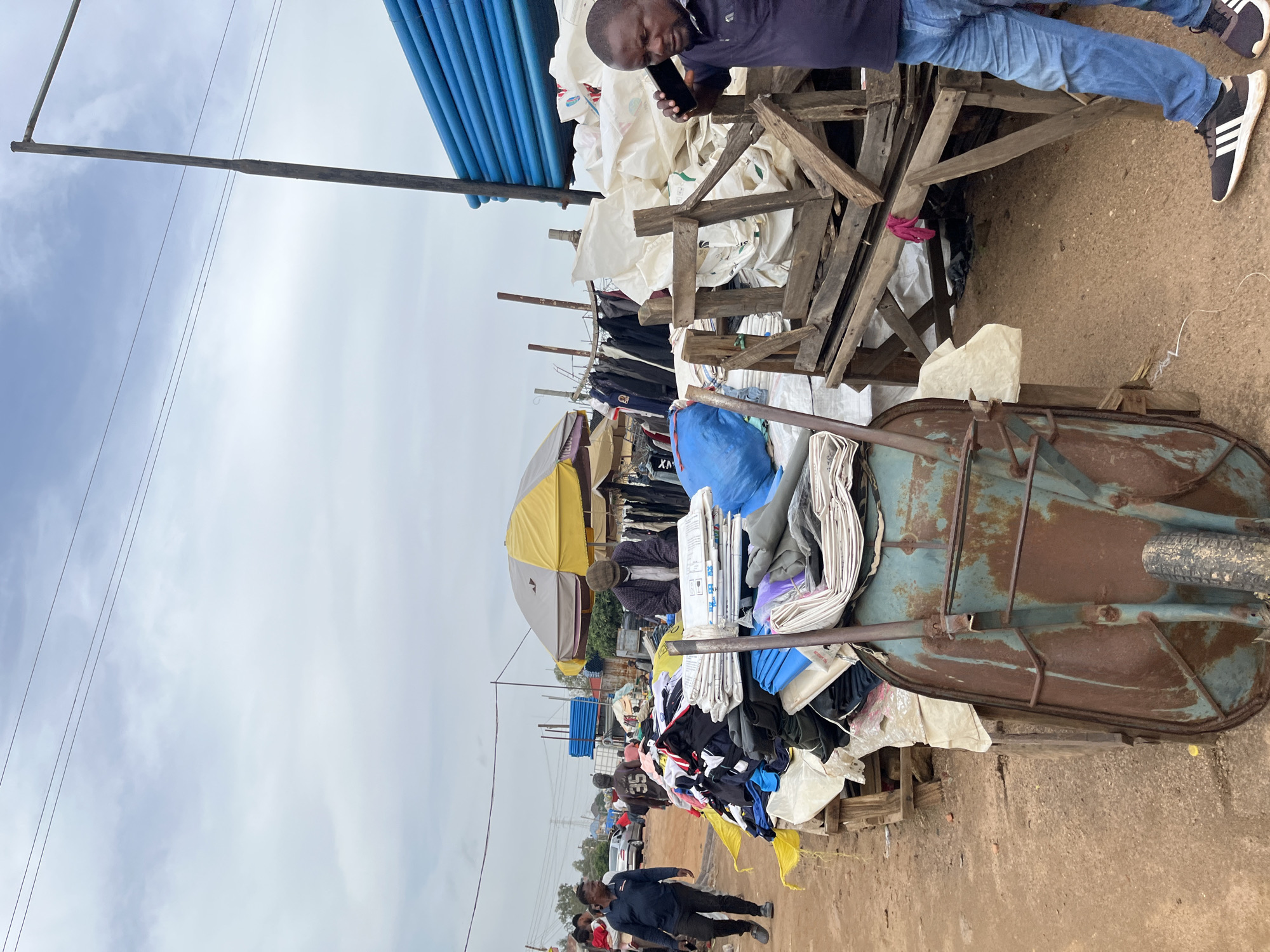 The media file depicts an outdoor market scene in Gura Top, Nigeria. The primary subjects are several people, including a man in the foreground wearing blue jeans and a dark long-sleeved shirt, looking at his phone. He is standing near a collection of items on a wooden structure.
The scene is cluttered with various objects, predominantly piles of clothing and fabrics, some in large sacks or bags. There is a rusty green wheelbarrow filled with what appears to be fabric or clothing. Wooden planks and beams are scattered throughout the scene, forming makeshift stands or supports for the goods. A yellow and grey umbrella is visible in the background, suggesting a marketplace setting. Blue plastic pipes are stacked in the upper left portion of the image.
The activity appears to be the operation of a market stall, with goods laid out for display or sale. The man in the foreground seems engaged with his phone, possibly a vendor or a customer. Other people are visible in the background, some appearing to be walking through the market.
Notable details include the cloudy sky, indicating overcast weather. The ground is a mix of dirt and sand. The overall atmosphere seems busy, typical of a market environment. There is no visible text in the media file. The time of day is likely daytime, given the lighting conditions.