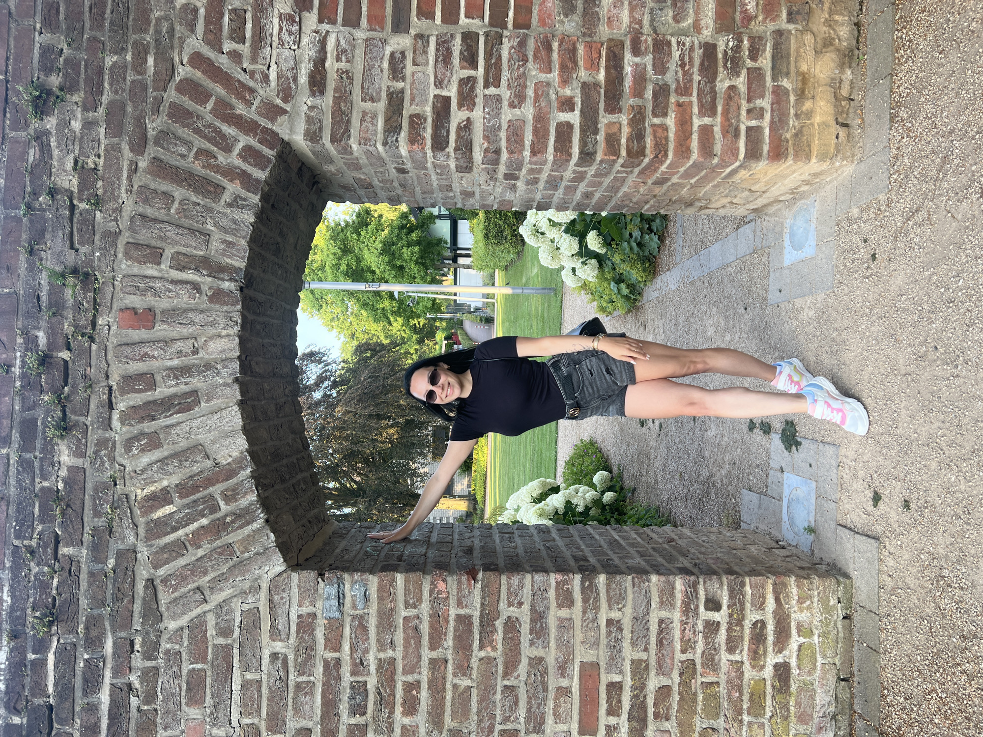 A full-body, eye-level, medium shot shows a young woman standing in a weathered brick archway. She is positioned in the center of the arch, facing forward, and smiling. Her right arm is extended and resting on the left pillar of the arch. She is wearing a black t-shirt, dark denim shorts, white sneakers with pink and blue accents, and sunglasses. A black purse is slung over her right shoulder. The archway is constructed from reddish-brown bricks and is partially covered in greenery. Beyond the arch, a pathway leads through a garden with white hydrangeas and lush green trees. The ground in the foreground and within the archway is covered in gravel and cobblestones. The scene appears to be in the gardens of Wassenberg Castle in Wassenberg, Germany.