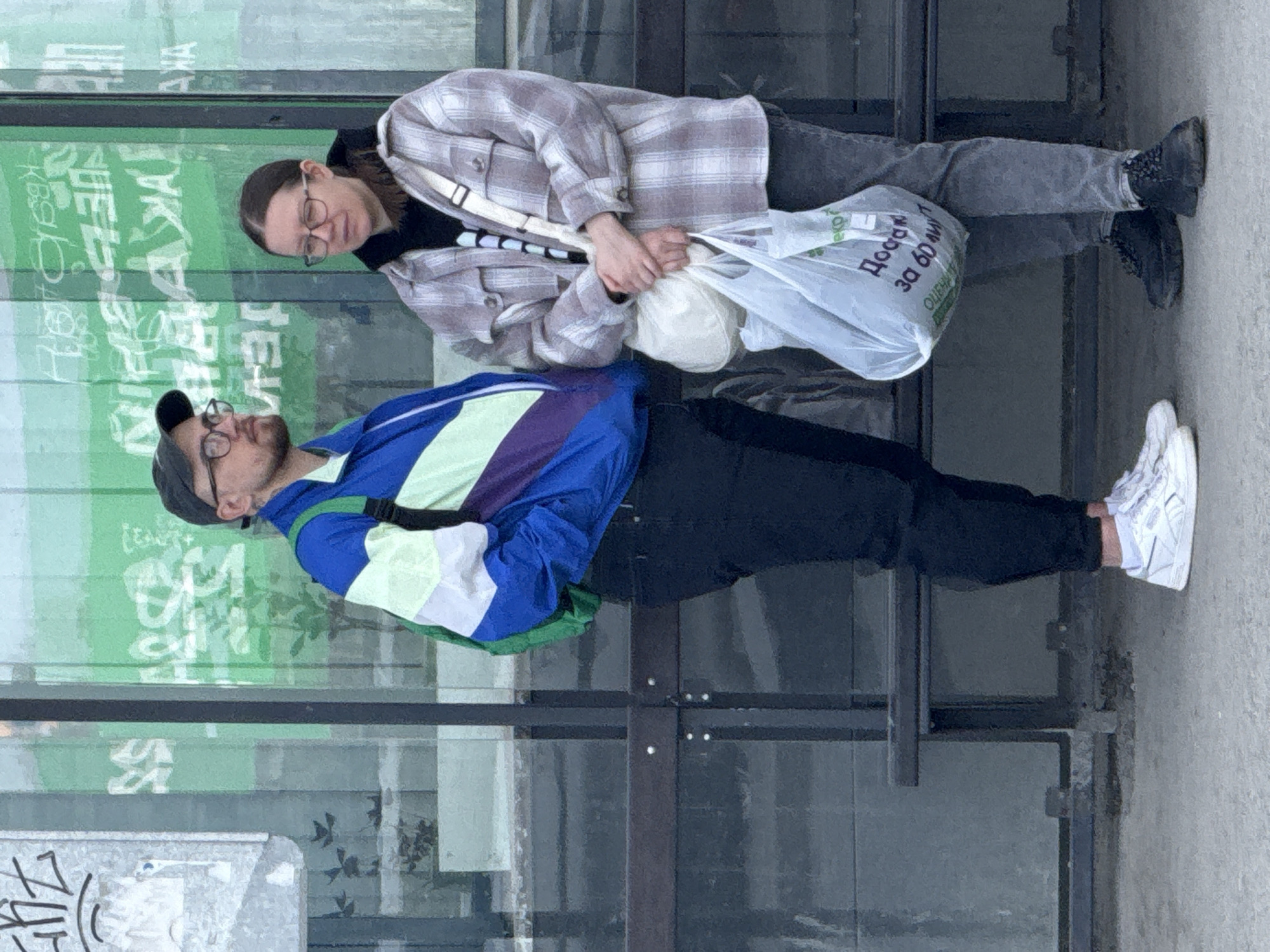 Two individuals are standing outdoors, likely in an urban setting, possibly in front of a building with large glass panels. The image is captured during the daytime with clear lighting.
To the left, a man is visible, wearing a dark cap, glasses, and a vibrant jacket featuring blocks of blue, green, purple, and white. He has dark pants, white sneakers, and a black backpack. His gaze is directed slightly upwards and to the right.
To the right, a woman stands, wearing glasses, a light-colored plaid shacket over a dark top, dark jeans, and black shoes. She has a white shoulder bag across her body and is holding two plastic bags in her hands. Her expression is somewhat neutral or contemplative.
The background consists of large glass surfaces, possibly windows or doors, featuring reflections of green elements and white Cyrillic text, some of which is illegible due to reflections and distortion. A dark metal frame divides these glass panels. On the semi-transparent plastic bag held by the woman, Russian text is clearly visible, reading "Доставка за 60 минут" (Delivery in 60 minutes) and "ЭКО" (ECO). Below this, "ОЦЕНКА" (Assessment/Rating) is partially discernible.
The ground appears to be concrete, and there's some graffiti on a lighter surface in the bottom left corner. The individuals seem to be standing or waiting together. The visible Russian text on the bag and reflections, combined with the provided location context, confirms the setting as Yekaterinburg, Russia.