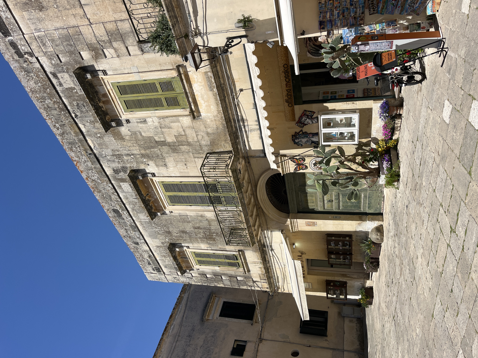 The image captures a street scene in Otranto, Italy, presenting a building with pale yellow facades and weathered stone. The building features green shuttered windows, wrought-iron balconies, and awnings above storefronts. A sign reads "officina mosaico," indicating a mosaic workshop. A bicycle is parked nearby. Postcard displays suggest a tourist-oriented business. The pavement is composed of light-colored stone blocks. The sky is a clear, deep blue. The scene suggests a sunny day in an old Italian town.