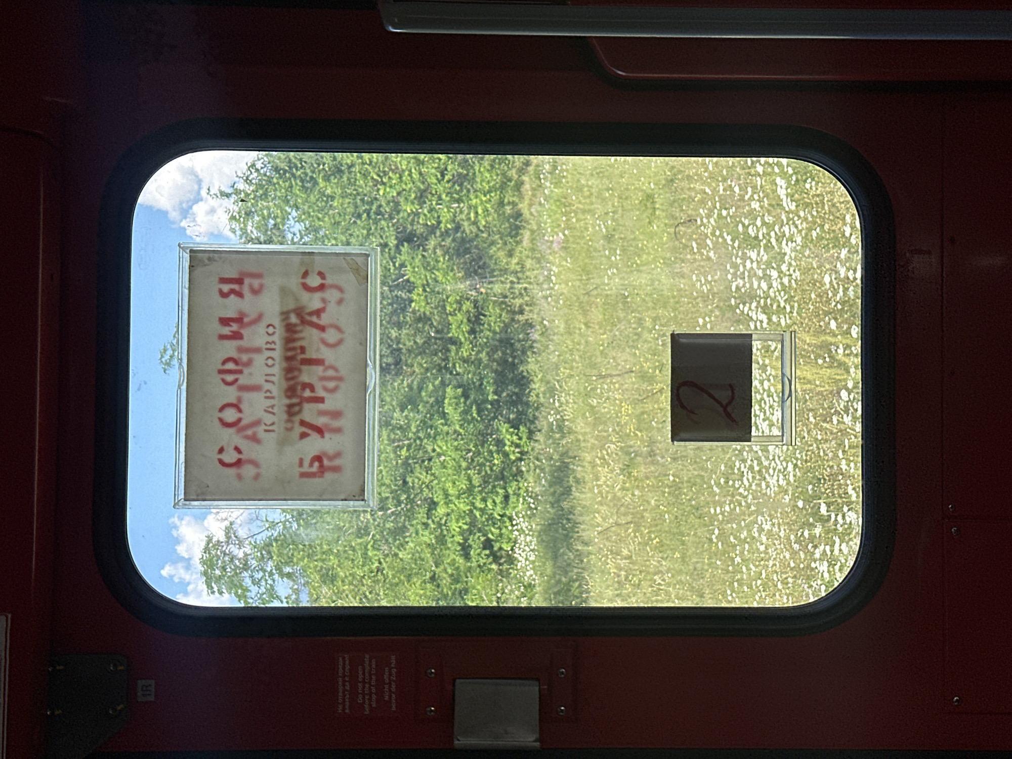 The image is an interior view from a train carriage window looking out at a field of wildflowers and trees under a blue sky with clouds. On the interior of the train door, there is a sign with Cyrillic text in red stencils. The text appears to read "СОФИЯ КАРЛОВО" on the top line and "БУРГАС" on the bottom line. These are likely destinations or routes. There are also smaller signs on the door with text in Bulgarian, English, and German stating "Do not open before the complete stop of the train" and "Nicht offnen bevor der Zug halt". A small sign with the number "2" is visible on the lower part of the window. The overall impression is that of a train carriage stopped or moving slowly through the Bulgarian countryside, near Karnare, a town known for its railway station.