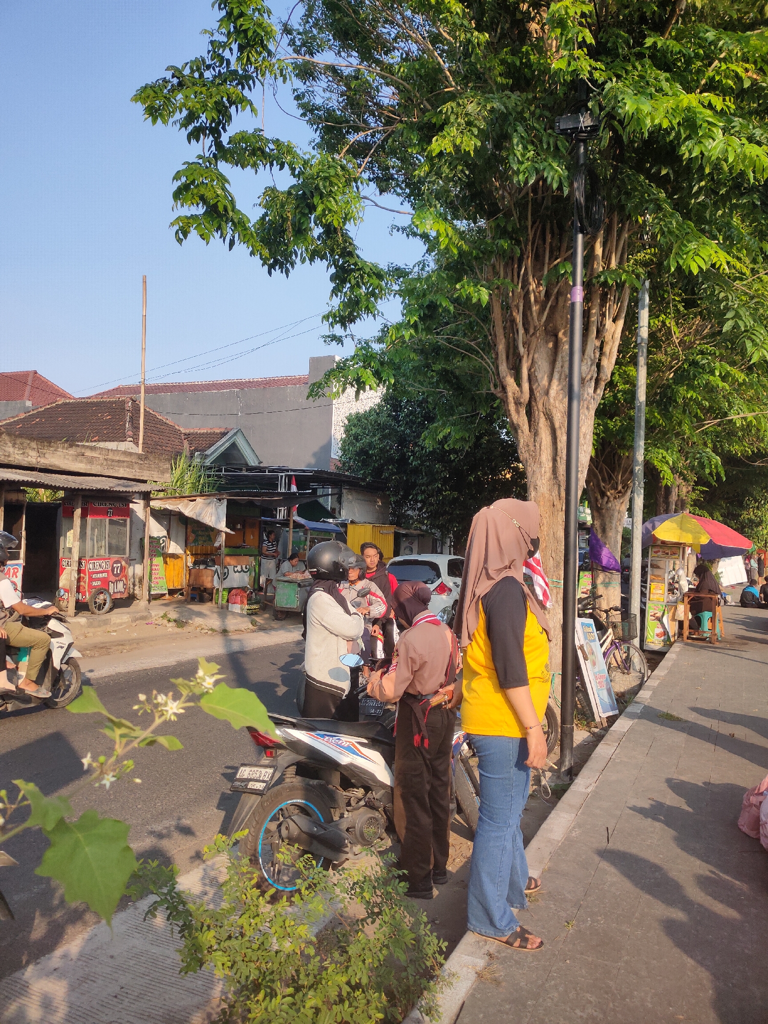 In Ngasem, Indonesia, a street scene unfolds with a mixture of daily life and commercial activity. A young woman wearing a hijab and a yellow t-shirt, coupled with blue jeans and sandals, stands on the sidewalk. Beside her, another individual in a scout uniform, a brown shirt and dark pants, is also present. The background reveals a row of food stalls, identified by signage like "CIRI ISI CIRENGISI 77" and various colorful awnings. Several motorcycles are parked along the street, with one in the foreground showing a license plate reading "AG 5852 RX". A white car is also visible in the mid-ground. Trees with dense green foliage line the street, casting shadows on the sidewalk. The sky is a clear blue, indicating fair weather. The overall impression is of a bustling urban environment, likely during daylight hours, capturing a slice of everyday life in a local Indonesian setting.