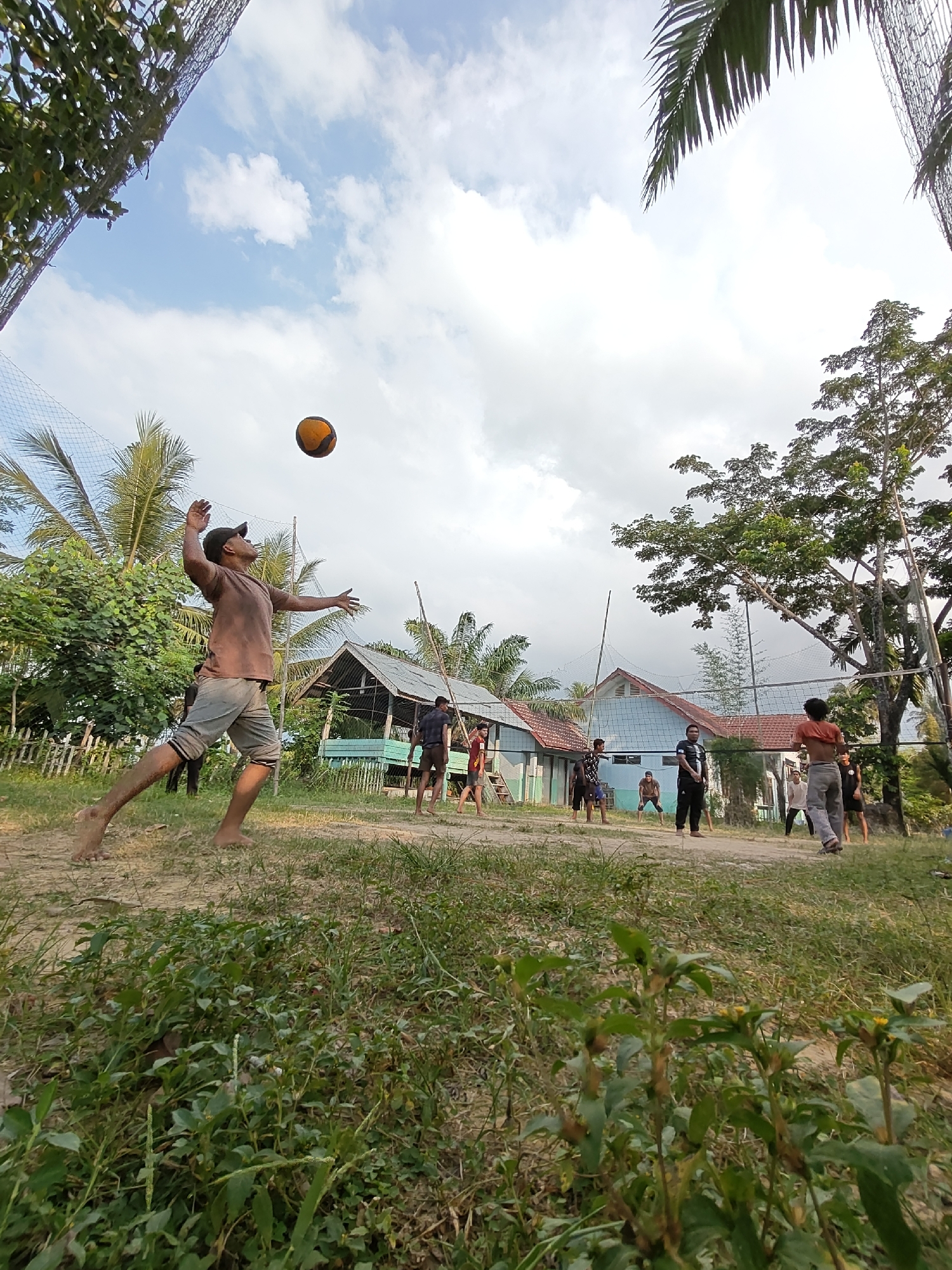 A group of young men are playing volleyball on a dirt court in Ubitpayaitek, Indonesia. The court is surrounded by lush greenery and palm trees, with a few simple buildings in the background. A man in the foreground, wearing a brown t-shirt and grey shorts, is in the process of serving the ball, his body contorted in mid-air as he reaches for the ball overhead. The ball is orange and yellow and suspended in the air. Other players are positioned on the court, some in ready stances, others observing the serve. The sky is a bright blue with scattered white clouds. A volleyball net is stretched across the court. The scene depicts a casual outdoor activity, likely a friendly game among locals.