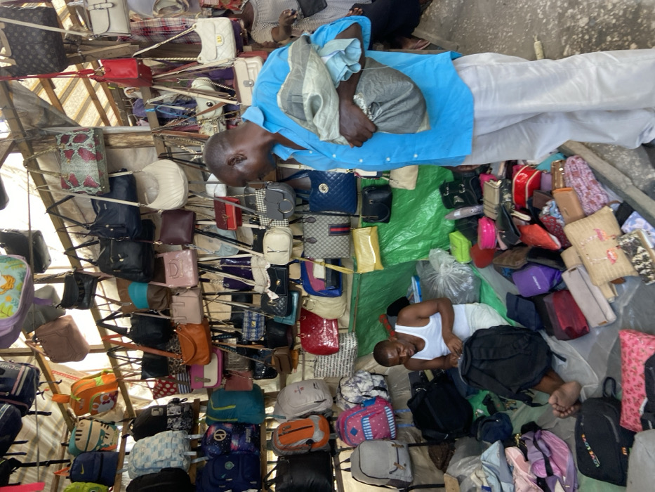 A rotated overhead shot shows a tightly packed market stall overflowing with handbags. Racks display diverse bags, colors, and styles. Two people are sleeping on the ground amongst the merchandise. One man wears a light blue shirt and lies on a light-colored cloth. Another man is in a white tank top. Bags and purses surround them on a green tarp.