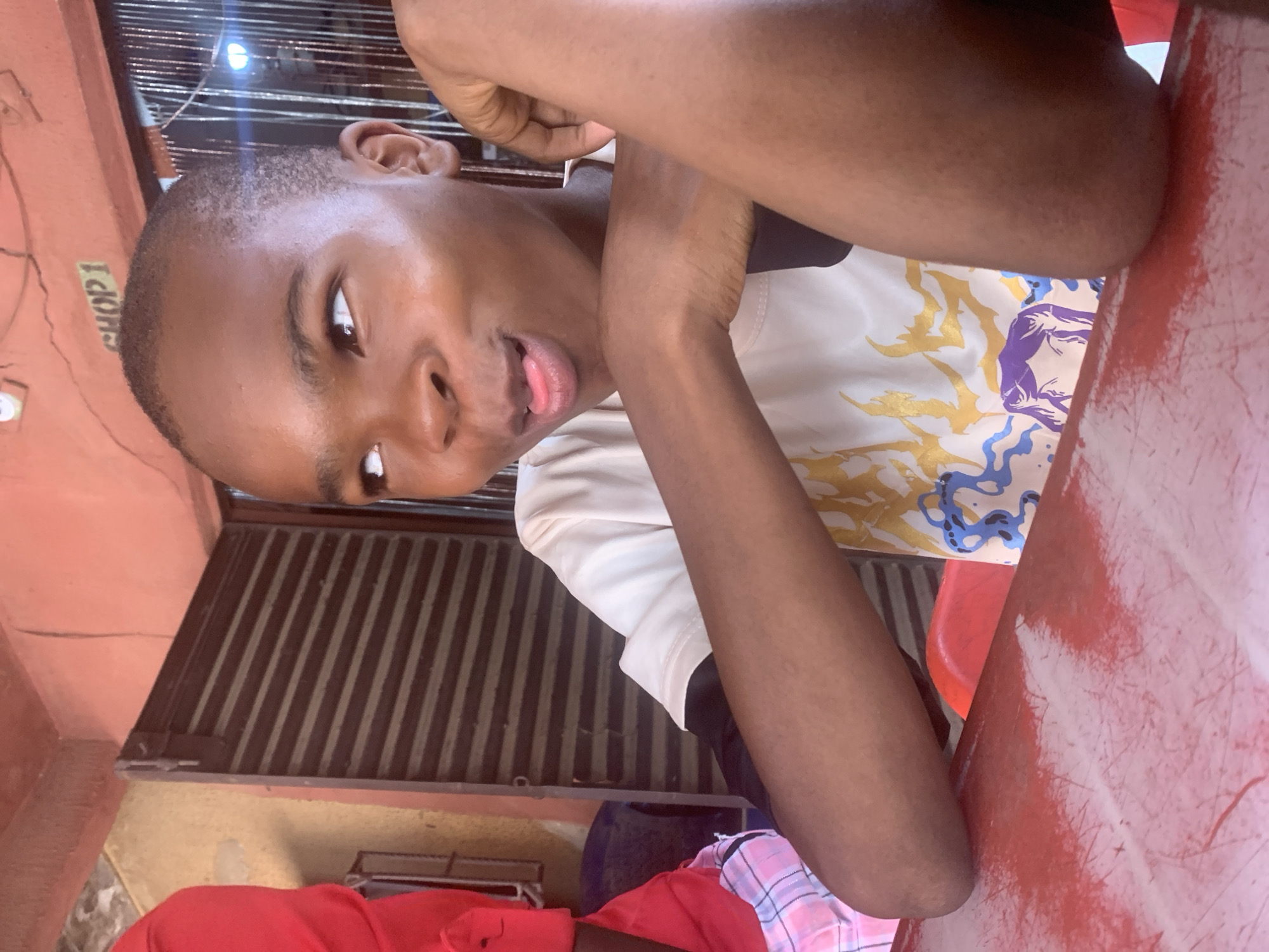 A young dark-skinned man is depicted from the shoulders up, leaning against a red table outdoors. He is wearing a short-sleeved shirt with a graphic print, and his arms are crossed in front of him. He is sticking his tongue out and looking to the side. A metal security gate is visible in the background, along with a weathered wall with the word "SHOP1" written on it. The location is Onitsha, Nigeria. Additional red clothing and a pink patterned shirt are partially visible in the lower left.
