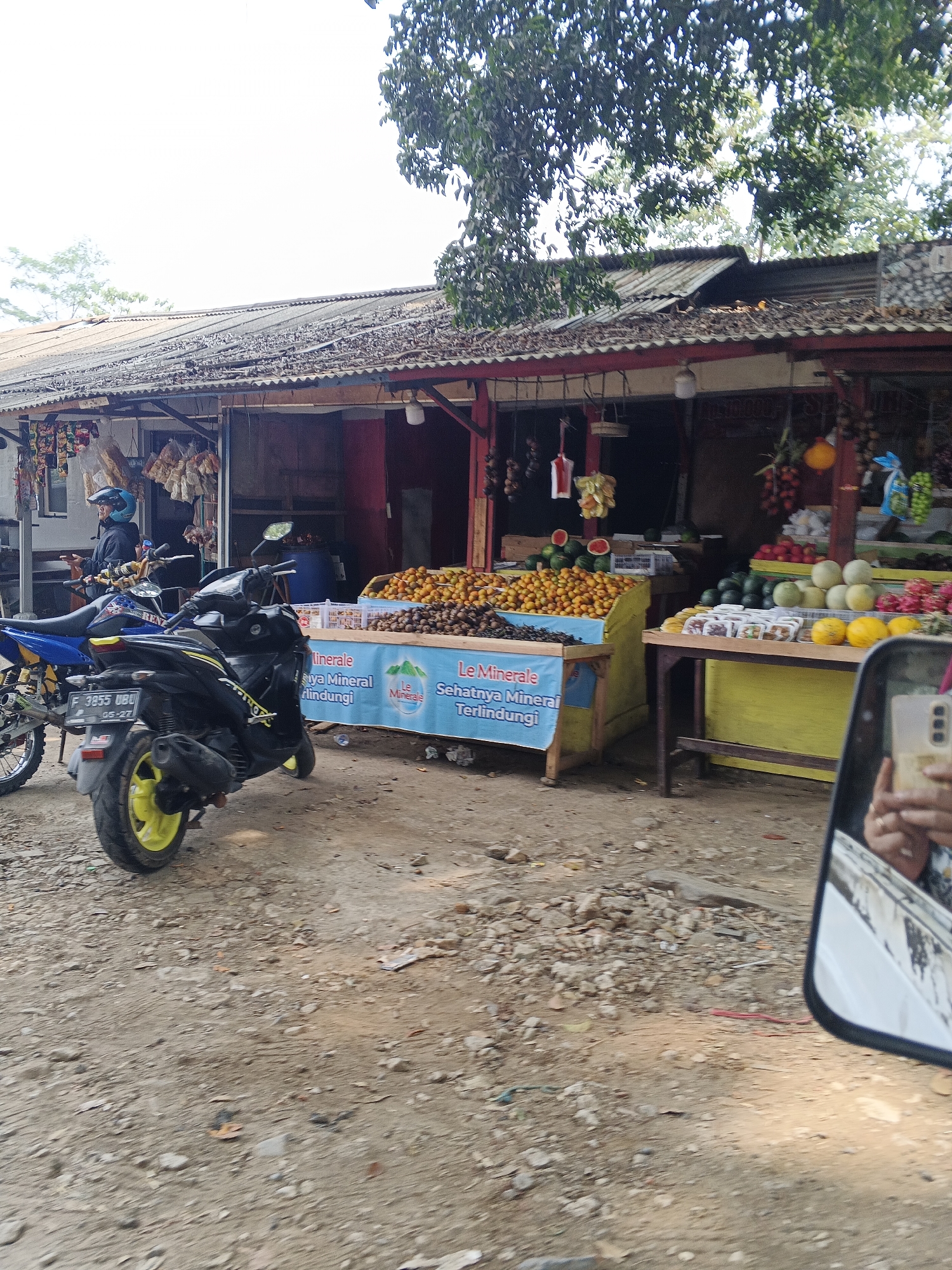 In Ubrug, Indonesia, a roadside market displays a variety of fruits and snacks. Two motorcycles are parked in front of the market stalls. The stall on the left displays packets of snacks and some dried goods. The main stall, in the center and right, is filled with an assortment of fruits including small orange fruits, watermelons, melons, and what appear to be grapes. A blue banner with Indonesian text is displayed across the front of this stall. A person wearing a blue helmet is seen on one of the motorcycles. The market is situated on a dirt road, with trees and foliage visible in the background. The scene is captured from the perspective of a vehicle, with a side mirror reflecting a person holding a mobile phone.