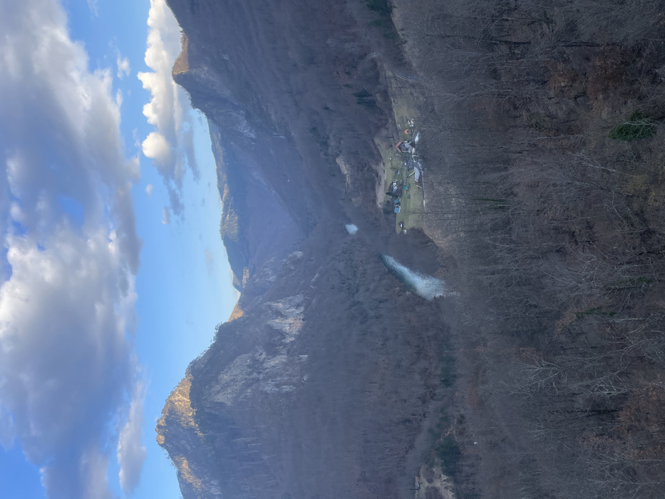 An aerial-perspective image captures a wide view of the Tara River Canyon in Đurđevića Tara, Montenegro, under a partly cloudy sky. The deep canyon, oriented generally from the upper center to the lower left, is dominated by steep, rugged mountainsides covered with bare deciduous trees, suggesting late autumn or winter. A turquoise-colored river, identified as the Tara River, snakes through the canyon floor. Sunlight illuminates the upper sections of the mountains, particularly a prominent peak on the left and ridges on the right, contrasting with the shadowed canyon depths. The sky is blue with scattered white and grey cumulus clouds. On the right bank of the river, in the mid-ground, a small settlement consisting of approximately seven structures with various roof colors, including orange and blue, is visible on a relatively flat area amidst the trees. The immediate foreground is comprised of the bare branches of trees, indicating a high vantage point. No individuals, animals, or specific actions are discernible.