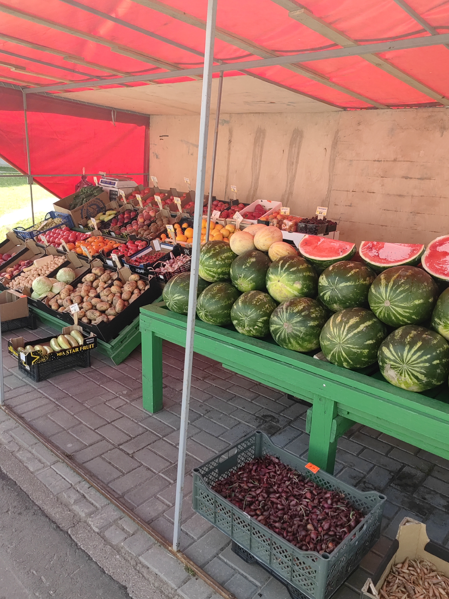 An outdoor fruit and vegetable stand displays a variety of fresh produce. The stand is covered by a red awning, supported by metal poles. On the left, crates of cucumbers and other vegetables are visible. In the center, a large display of potatoes is prominently featured, alongside cabbages and other root vegetables. To the right, watermelons are arranged on a bright green wooden table, with sliced watermelons placed on top. Crates filled with small red onions and what appear to be shallots are positioned on the ground below the table. The ground is paved with brick-like pavers. The overall scene suggests a bustling market environment.