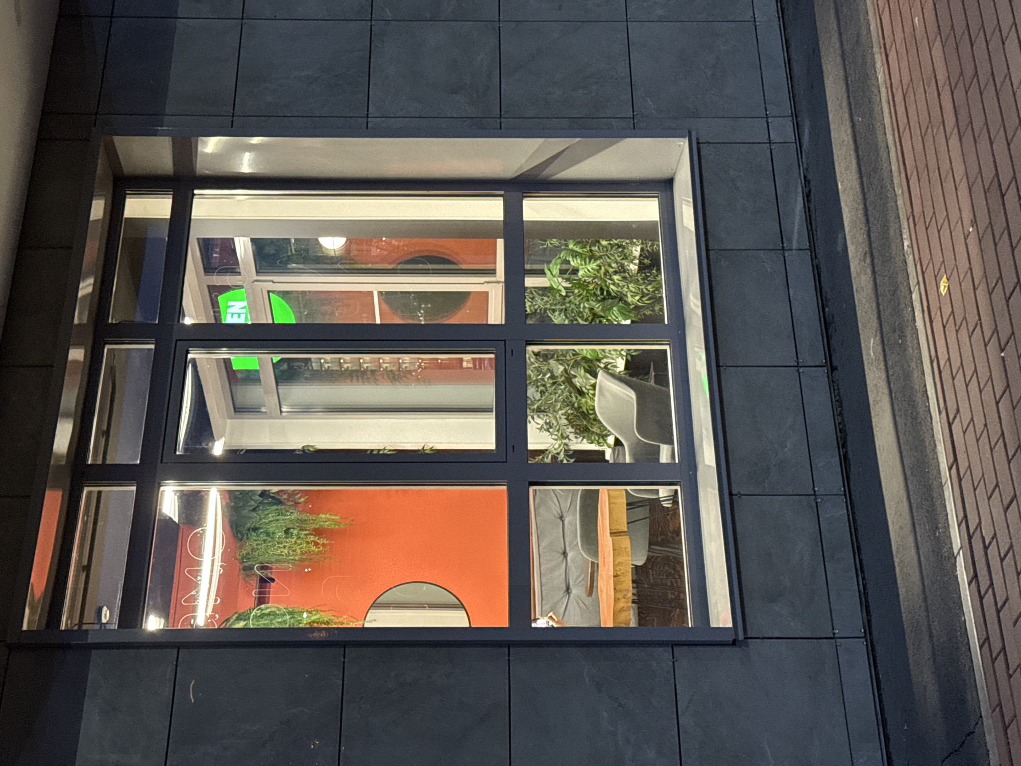 A window, viewed from outdoors, is mounted on a building wall composed of dark grey square tiles. The window itself is divided into six panes by black frames. The reflection inside the window suggests an interior scene with elements like greenery, furniture, and an orange wall with a half-circle cutout. The street/sidewalk with brick-like material is visible on the right side of the image. A possible sign with green lettering can be seen in the top left window pane. The image does not include enough information to confirm its location in Krasnoyarsk, Russia.