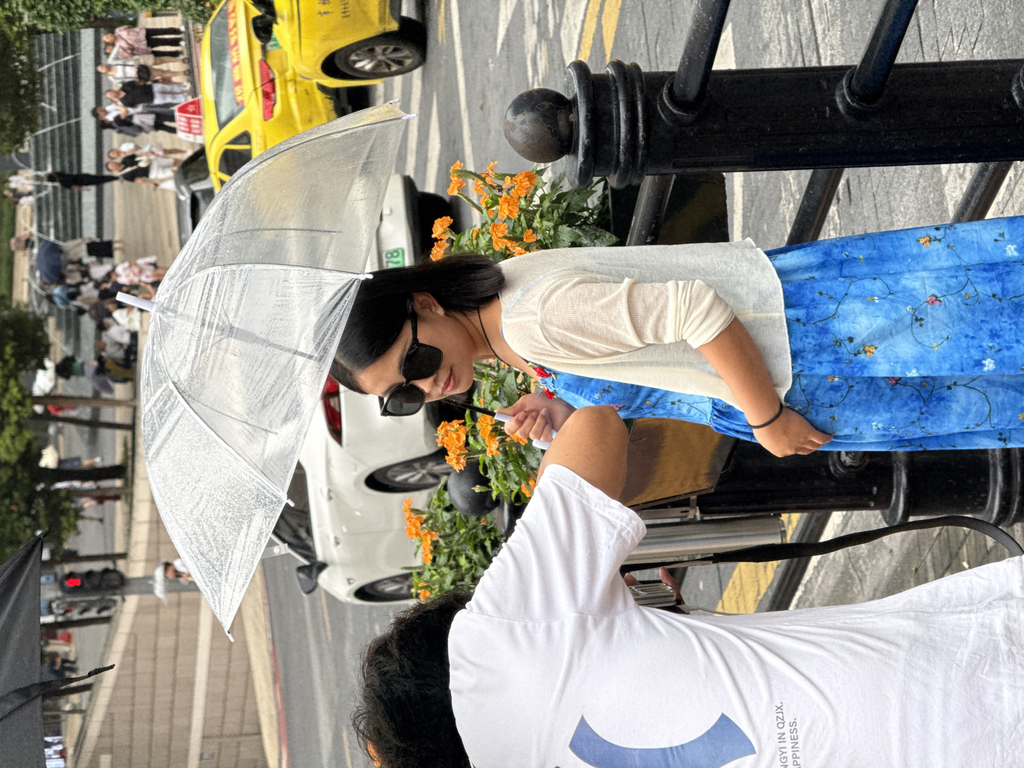 In Yuzhong District, China, during a rainy day, a young woman stands under a clear, water-covered umbrella. She is wearing a blue dress adorned with a floral pattern and a light-colored cardigan. Her face is partially obscured by large black sunglasses. To her left, a person wearing a white t-shirt with text that reads "NGY IN QZJX. HAPPINESS." is partially visible, holding a camera. In the background, yellow taxis and a white car are seen on the wet street, alongside a traffic light. Orange flowers are planted in a black planter, and a black metal railing with decorative posts is in the foreground. The scene is outdoors, with a blurred urban backdrop.