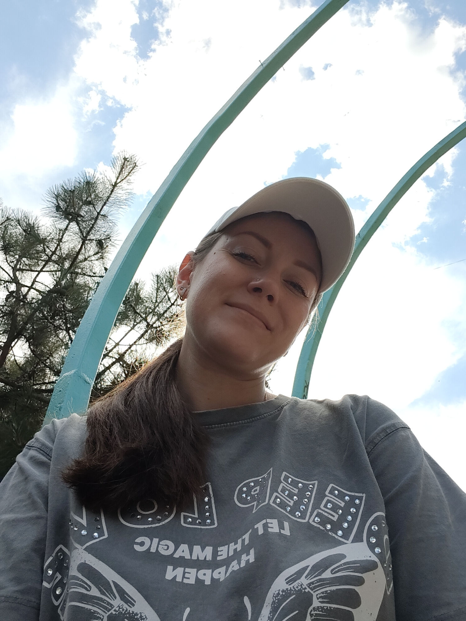A young woman wearing a grey t-shirt and a white baseball cap takes a selfie outdoors. She has her hair in a ponytail and is smiling. She is positioned under a light blue archway. In the background, there is a tree and a cloudy sky. The image appears to have been taken during the daytime. The location is unknown.