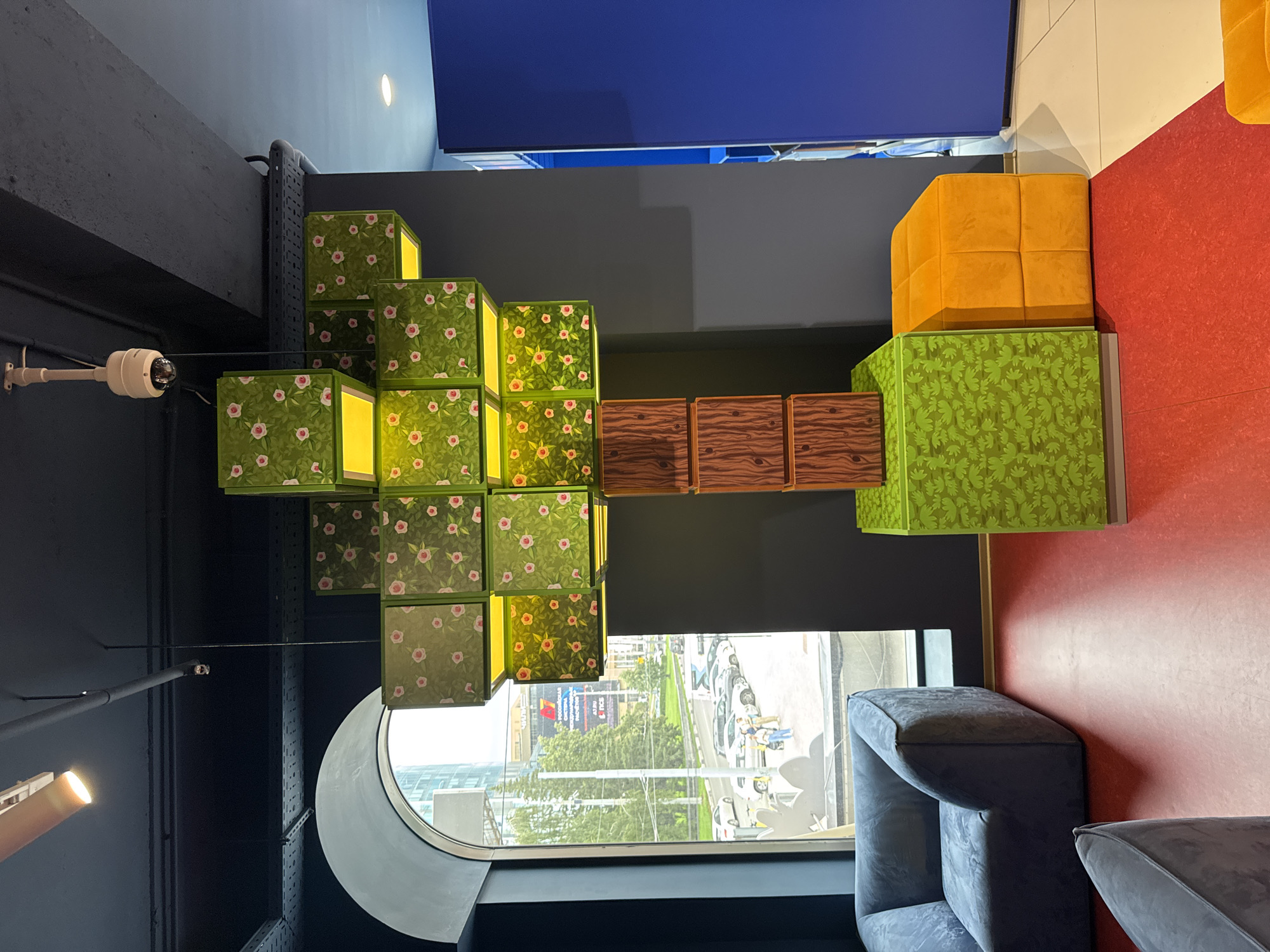 The image captures an indoor scene with a window view of an urban environment, likely Yekaterinburg, Russia. The interior features decorative, cube-shaped lamps with green floral patterns and yellow interiors, arranged in a tiered structure. To the right, there are two large, soft orange and green cubes, possibly seating or decorative elements. A grey, upholstered sofa is visible in the foreground, positioned next to a large, arched window. Outside the window, a street scene unfolds with cars and pedestrians, suggesting daytime. The lighting is somewhat dim, indicating an overcast day or an indoor setting with artificial lighting. The overall ambiance is modern and playful, possibly a lounge or public space. There is no visible text in the image.