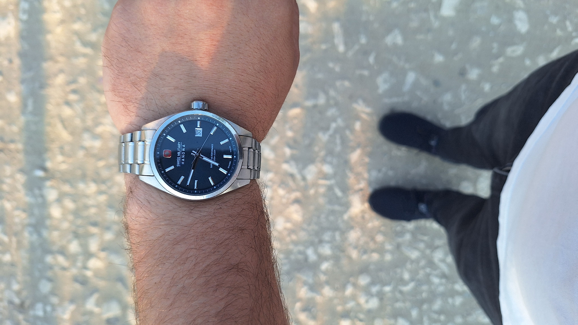 The image shows a close-up of a person's wrist wearing a Swiss Military Hanowa watch. The watch has a black face with silver indices and a date window displaying "30". The watch band is silver metal. The person's arm is hairy and is resting on a textured surface that appears to be gravel or small stones. In the background, the lower legs and feet of the person are visible, wearing black pants and black shoes. The ground is covered in the same gravelly texture. The image was taken in Sărata-Galbenă, Moldova.