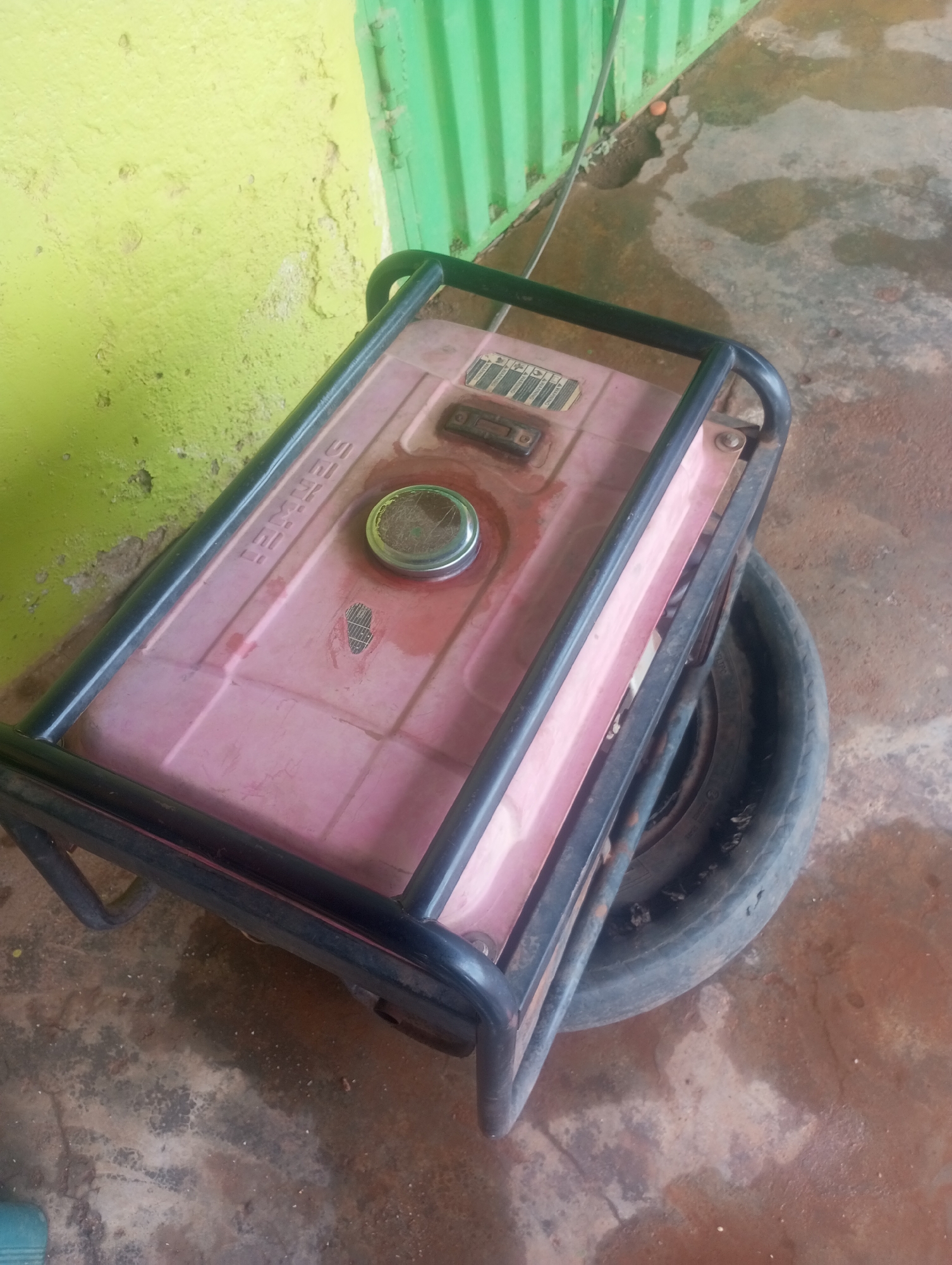 The image shows a red portable generator placed on the ground. The generator has a metal frame around it, and a black tire is visible underneath. The ground appears to be concrete and is wet in some areas. In the background, there is a lime green wall with some peeling paint and a corrugated green metal structure. The scene is outdoors, and the lighting suggests it is daytime. There are no people visible in the image.