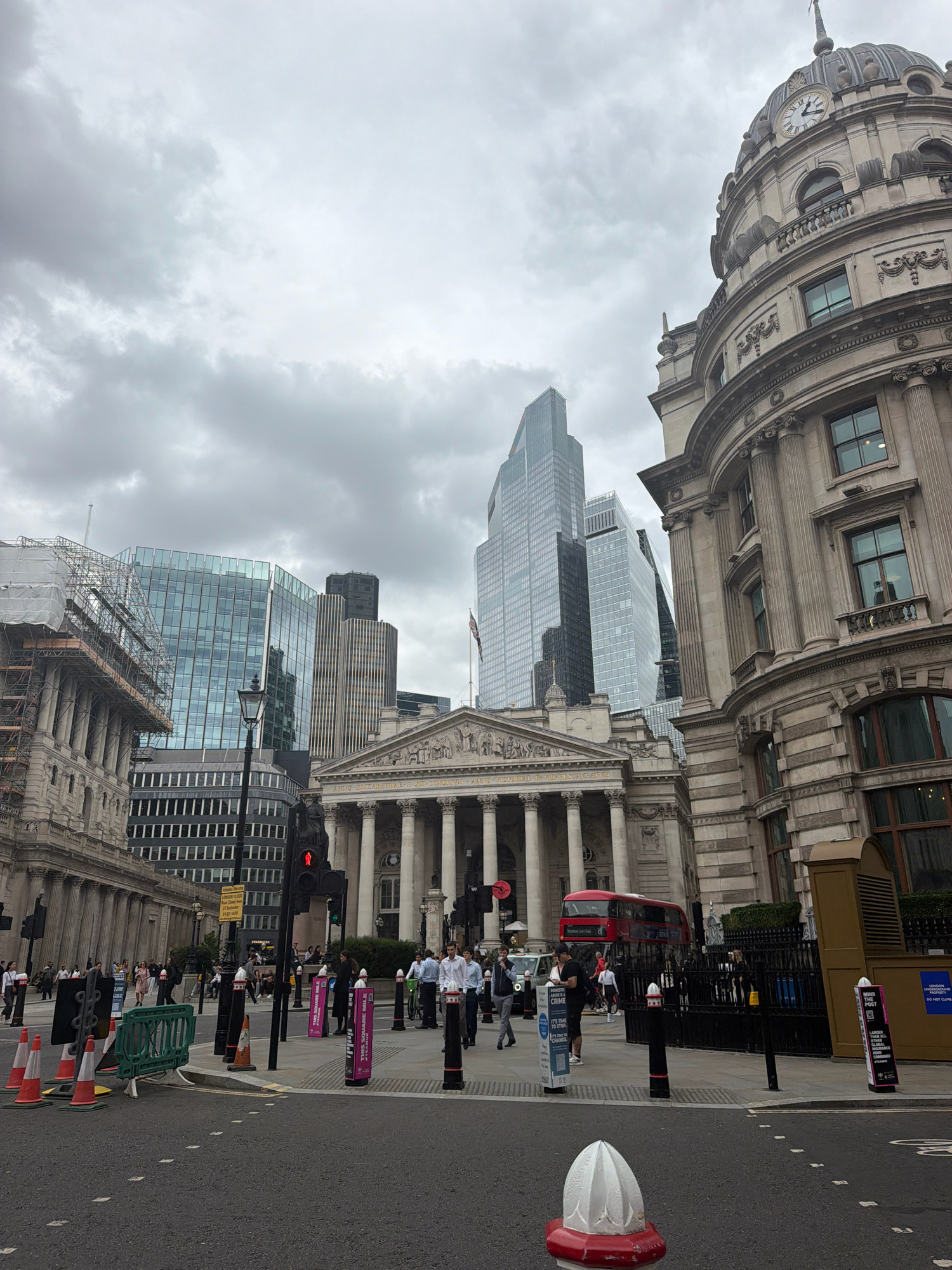 The image captures a street scene in the City of London, United Kingdom, characterized by a juxtaposition of historic architecture and modern skyscrapers under a cloudy sky. In the foreground, a street intersection is visible with traffic cones, bollards, and road markings. A red double-decker bus is crossing the intersection, and pedestrians are walking on the sidewalks.
To the right, a grand, ornate building with a dome and a clock face dominates the frame, exhibiting classical architectural features like columns and intricate carvings. To the left and in the background, modern glass-fronted skyscrapers rise into the sky, reflecting the overcast conditions. One of these modern buildings appears to be under construction or renovation, with scaffolding and protective sheeting visible.
Several people are scattered throughout the scene, some walking, others seemingly waiting or observing. The overall atmosphere is that of a bustling urban environment, typical of London's financial district. A traffic light shows a red signal for pedestrians, indicating a stop. Signs with various text and graphics are present, including one that appears to be an advertisement or information board. The ground is paved with asphalt and marked with white lines. The sky is uniformly gray and overcast.